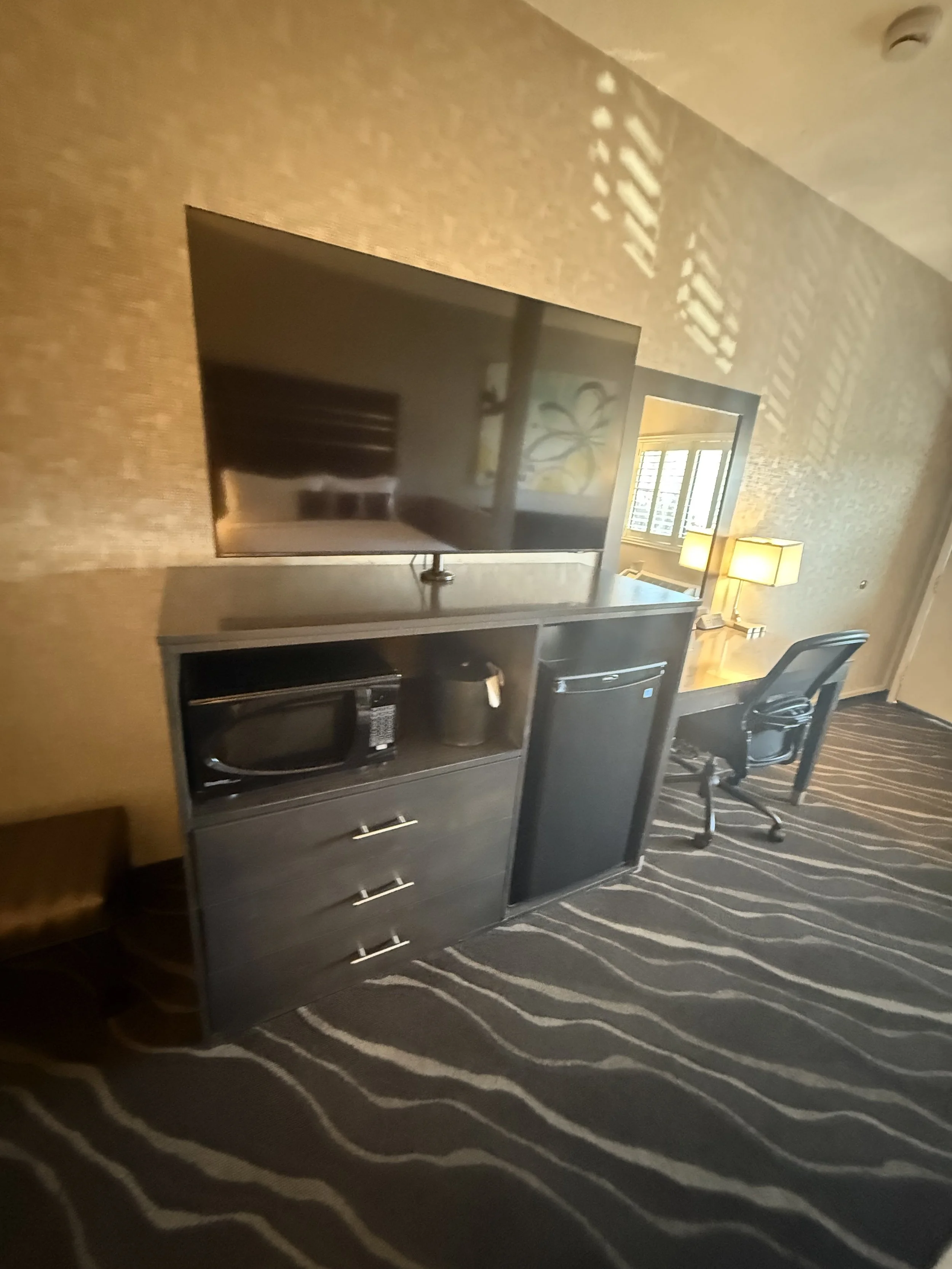A hotel room with a flat-screen TV on a black dresser, a microwave oven, a mini refrigerator, and a work desk with a black office chair and two lamps.