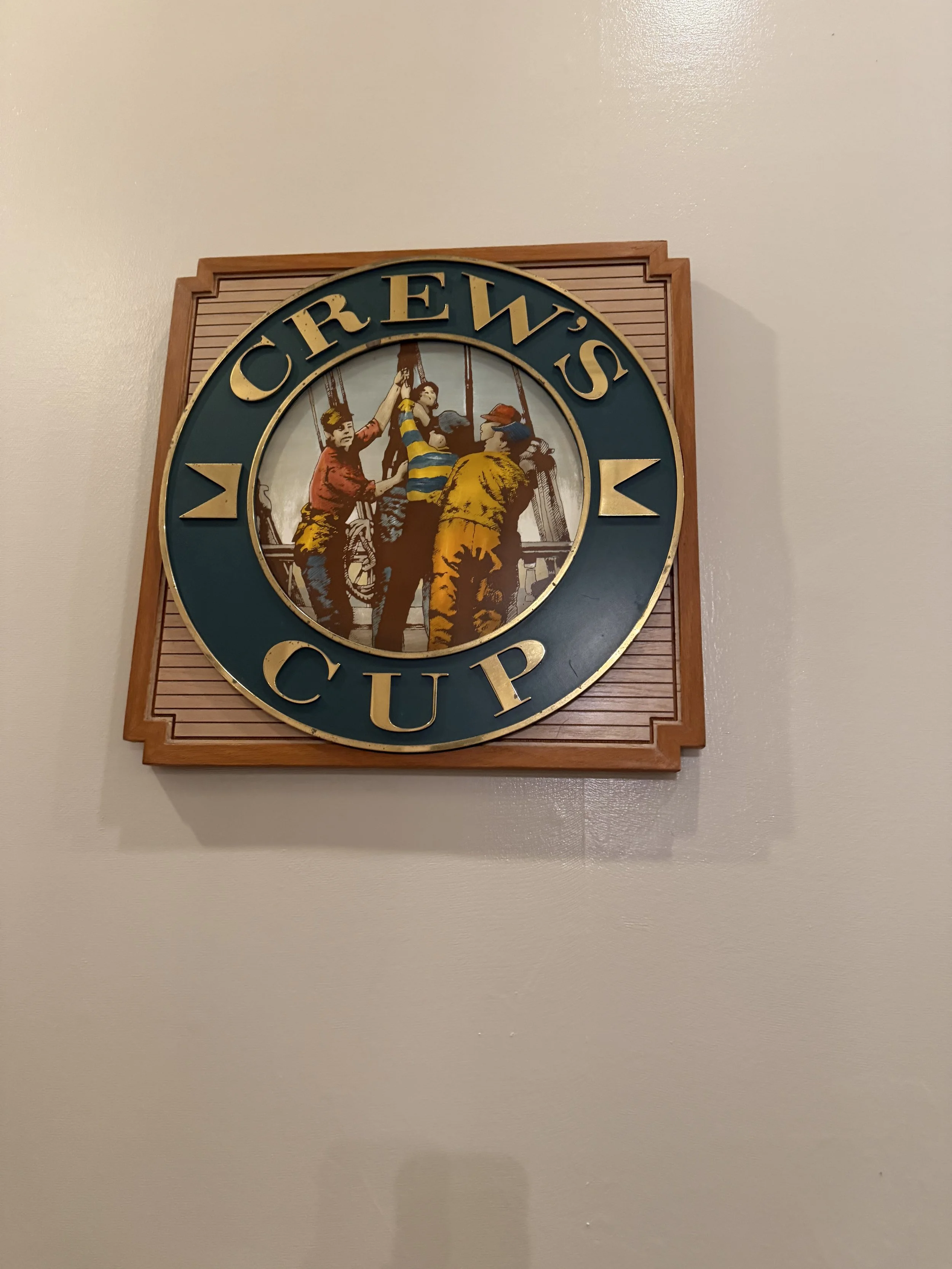 Decorative sign with the words 'Crew's Cup' and an image of three people, one of whom is holding a child in a striped shirt, on a wall.