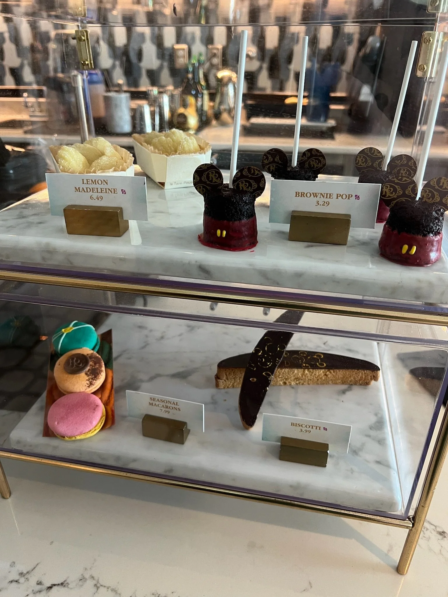 A display case with various desserts, including lemon madeleine, brownie pop, seasonal macarons, and biscotti on a marble surface.