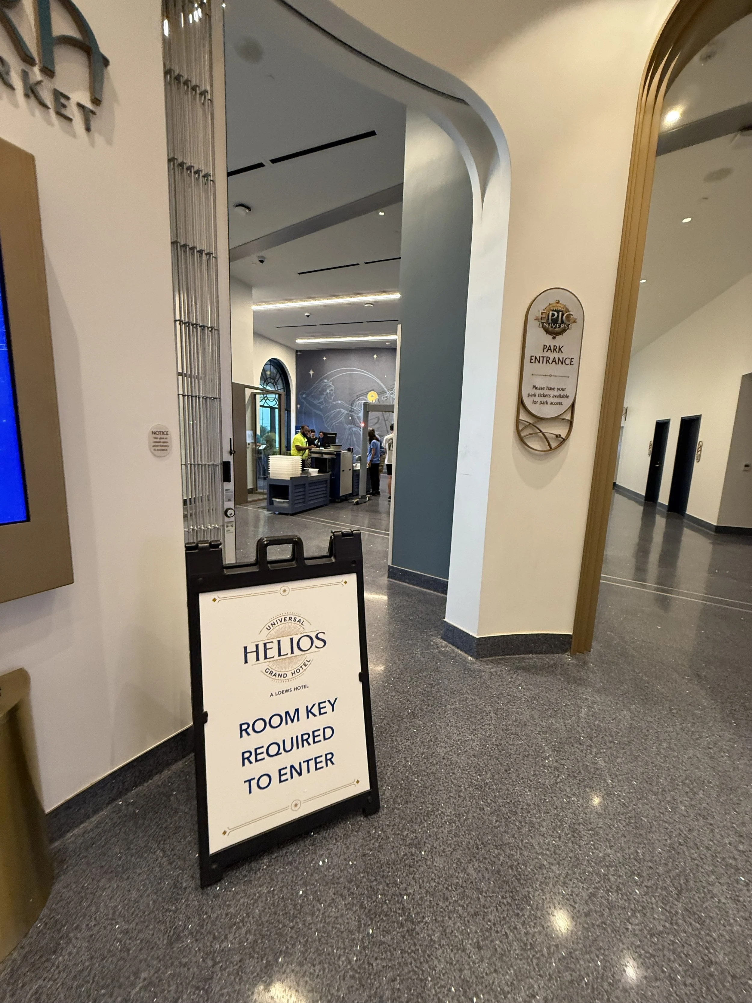 Hotel lobby area with a sign that reads 'Room key required to enter' in front of a doorway leading to a reception desk, with several people working behind the desk and a decorative wall with a space-themed design.