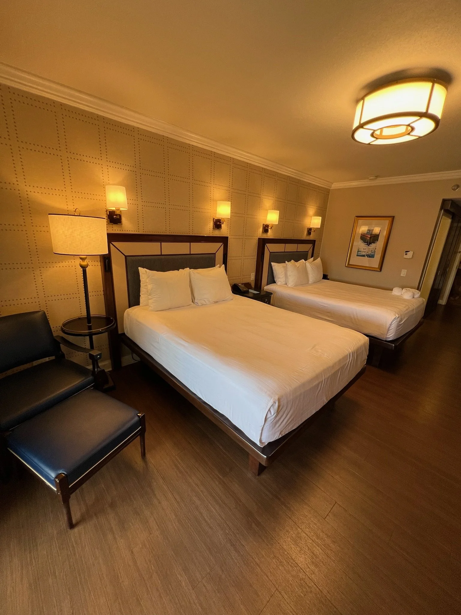 Hotel room with two beds, a lamp, chairs, wall-mounted lights, and framed artwork.