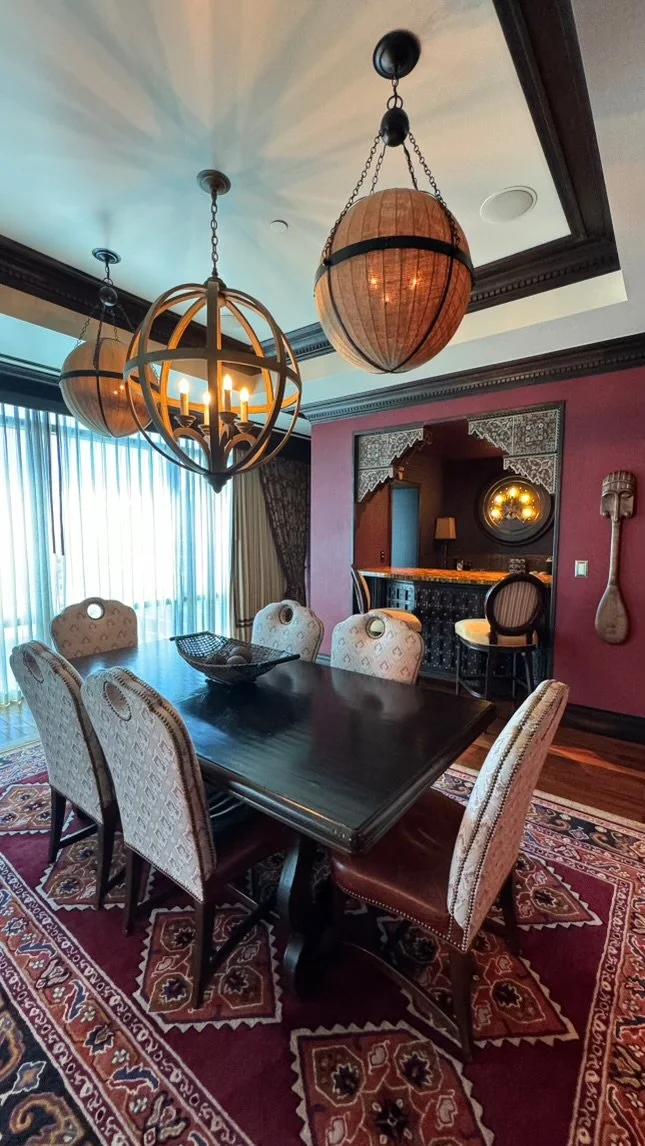 A dining area with a dark wooden table, six upholstered chairs, a patterned rug, and a hallway with a bar area in the background. There are three hanging lamps with wooden and metal designs, and a large window with curtains lightens the room.