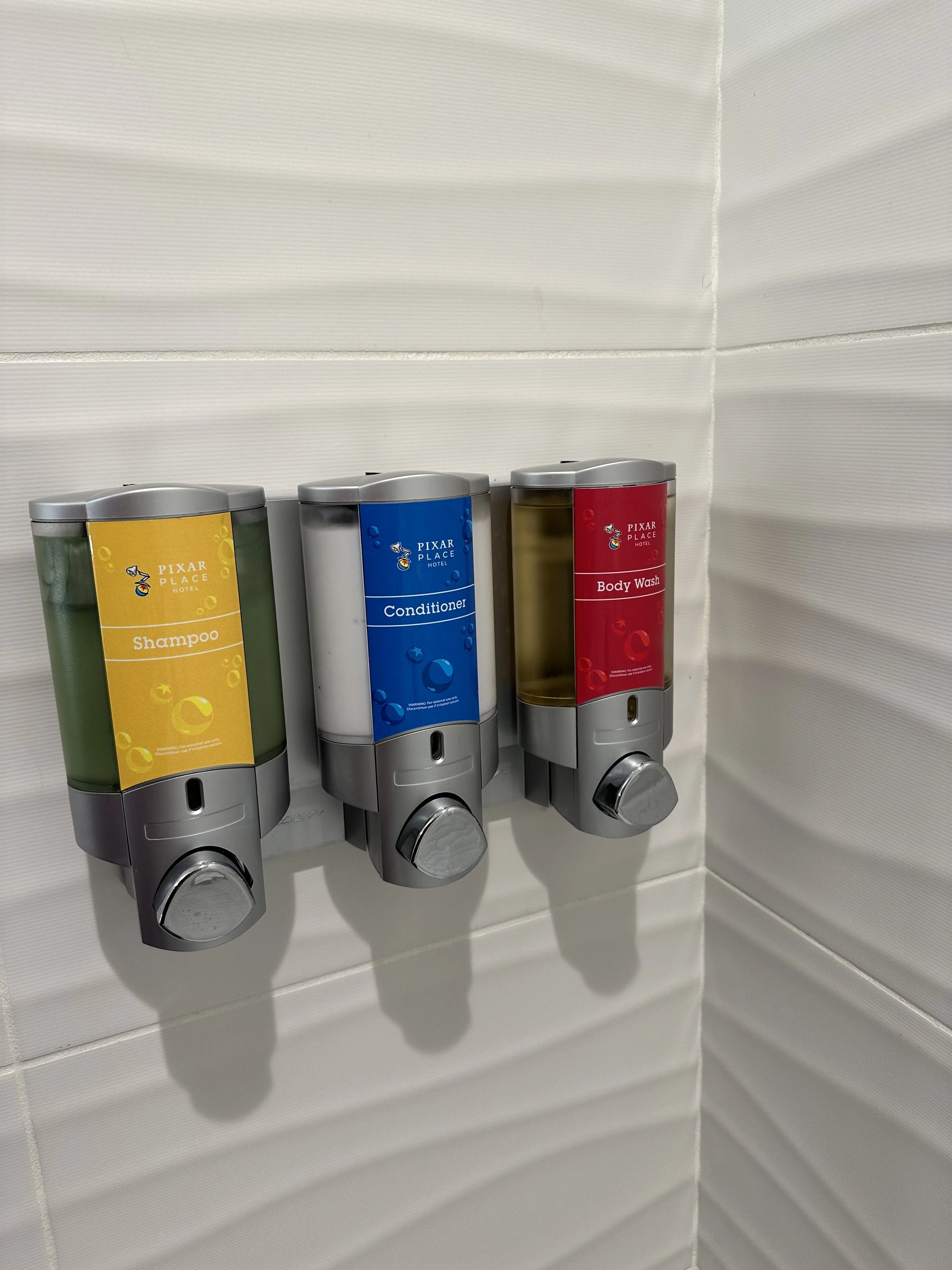 Three wall-mounted soap and shampoo dispensers labeled for shampoo, conditioner, and body wash, mounted on creamy white tiled wall.