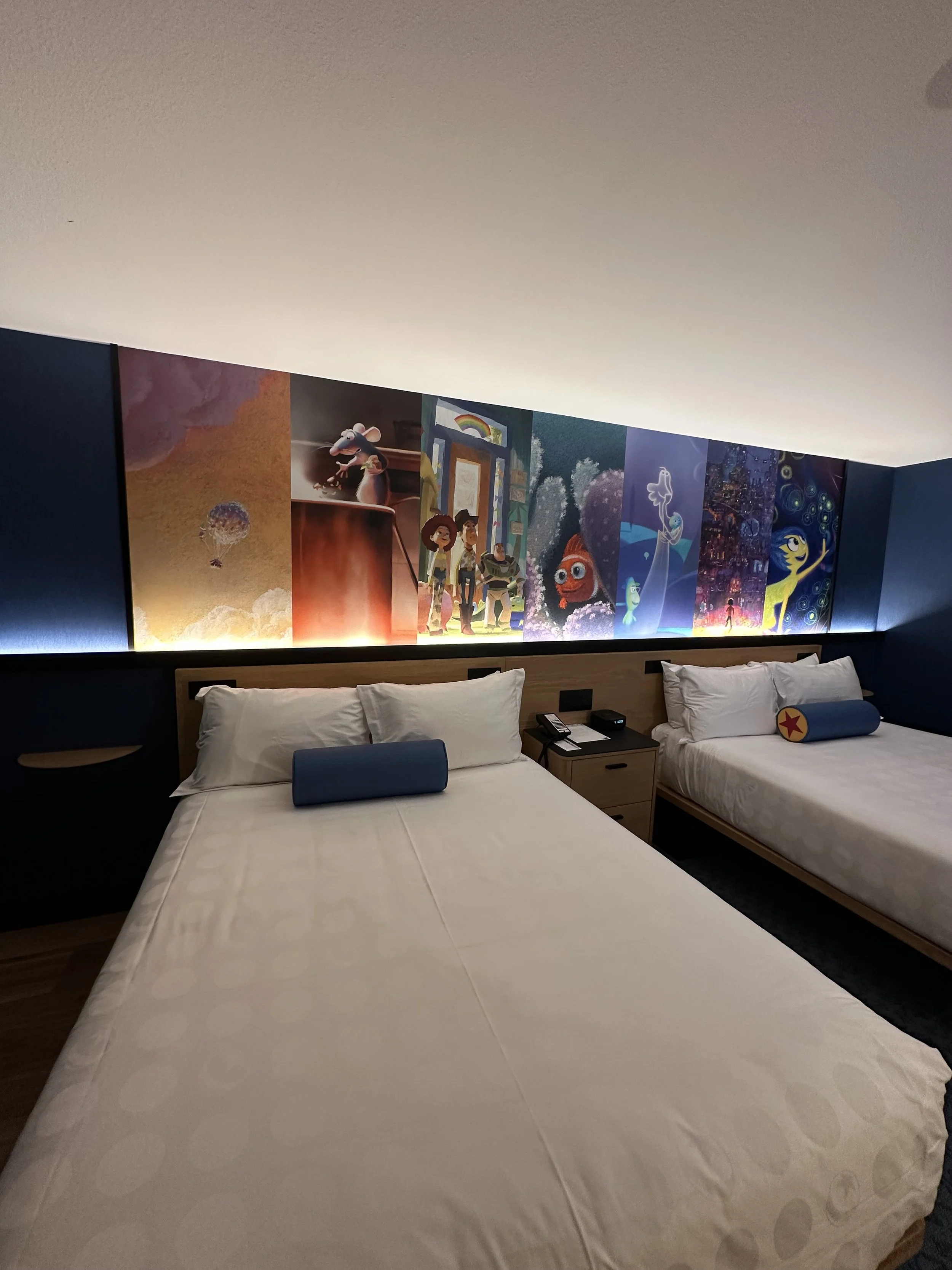 Hotel room with two beds, each with a burlap pillow and a blue bolster. A colorful mural of animated characters runs across the wall behind the beds. There is a nightstand with a telephone, remote, and alarm clock between the beds.