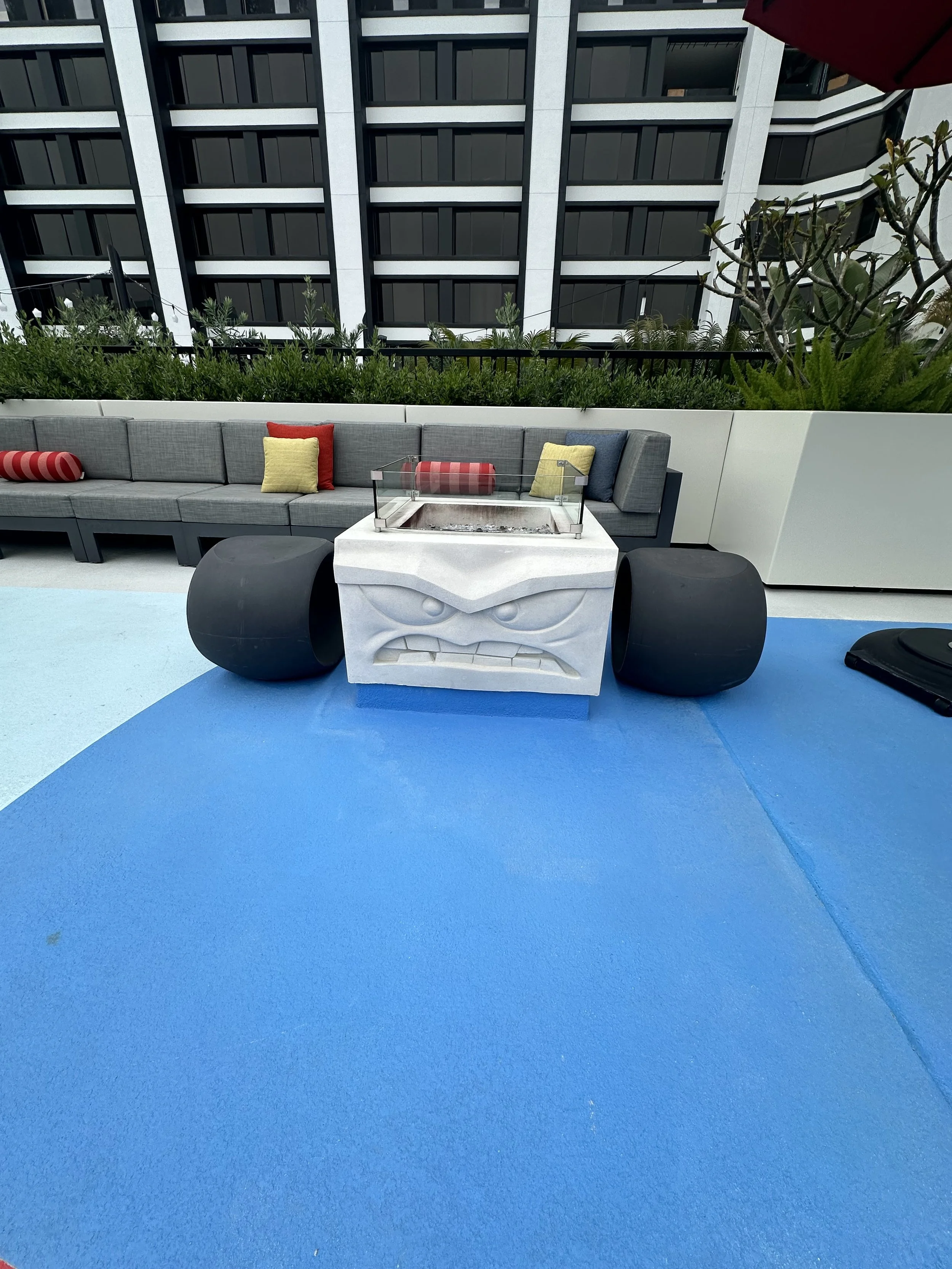 Outdoor seating area with a long gray sectional sofa with red, yellow, and blue cushions, greenery in the background, and a balancing platform with a carved face design and large black wheels in front of the sofa. Blue flooring surrounds the setup.