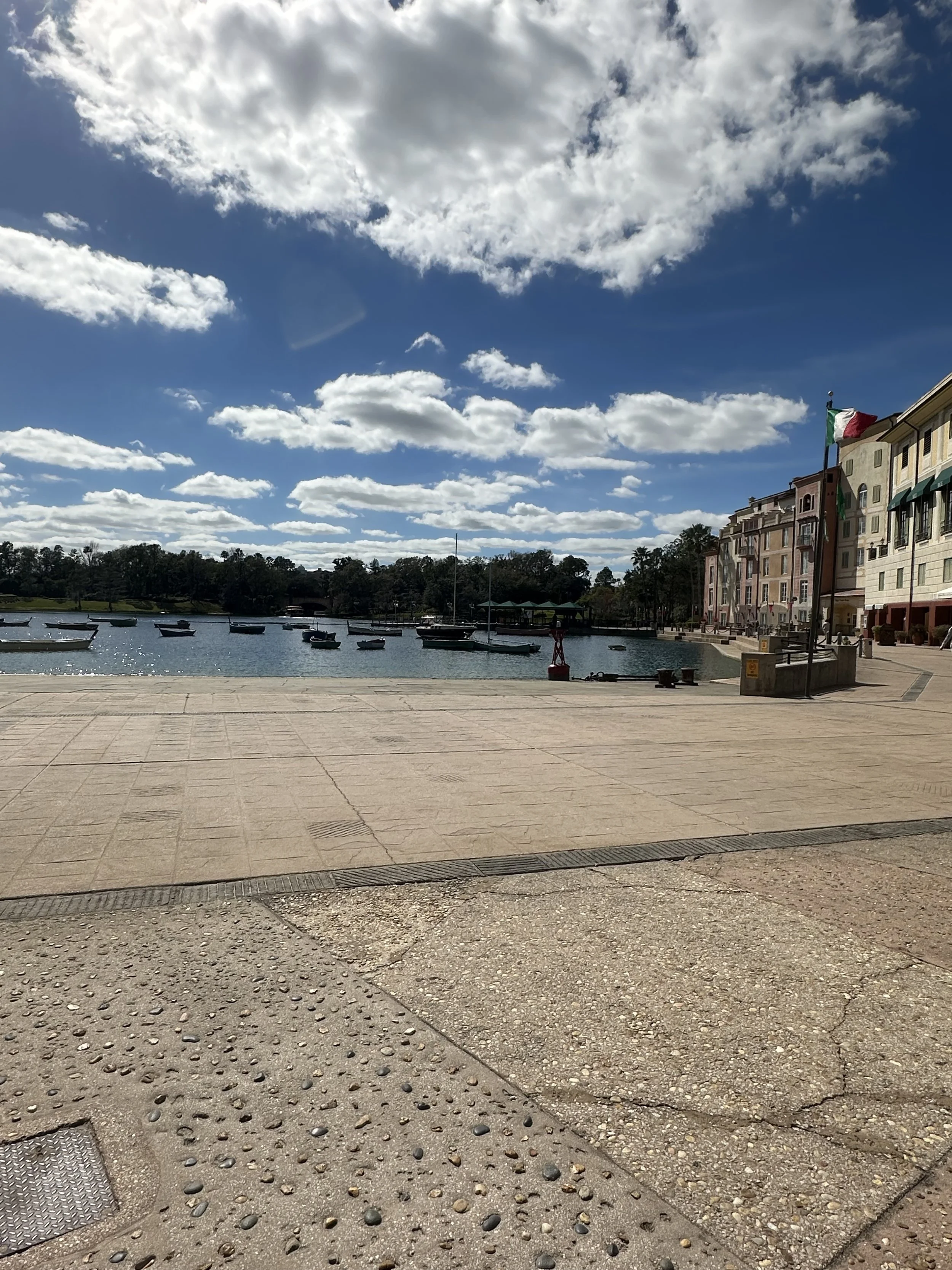 A waterfront scene with sailboats docked on a bay, colorful multi-story buildings with balconies and awnings, flags flying, and a paved promenade under a partly cloudy blue sky.