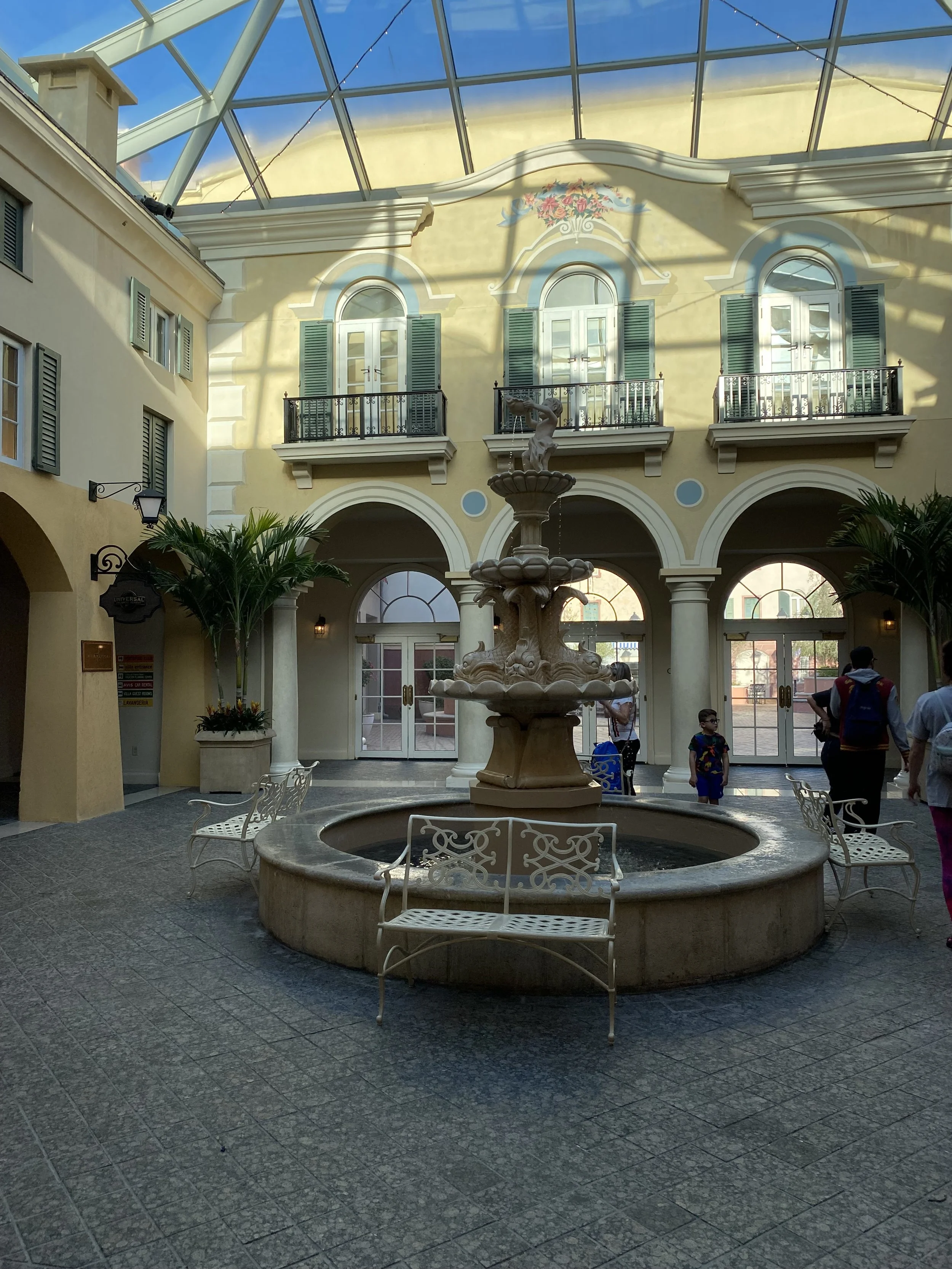 Indoor courtyard with a fountain at the center, surrounded by white benches, potted plants, and a yellow building with green windows and sliding doors. There are children and adults walking nearby, and a glass ceiling overhead.