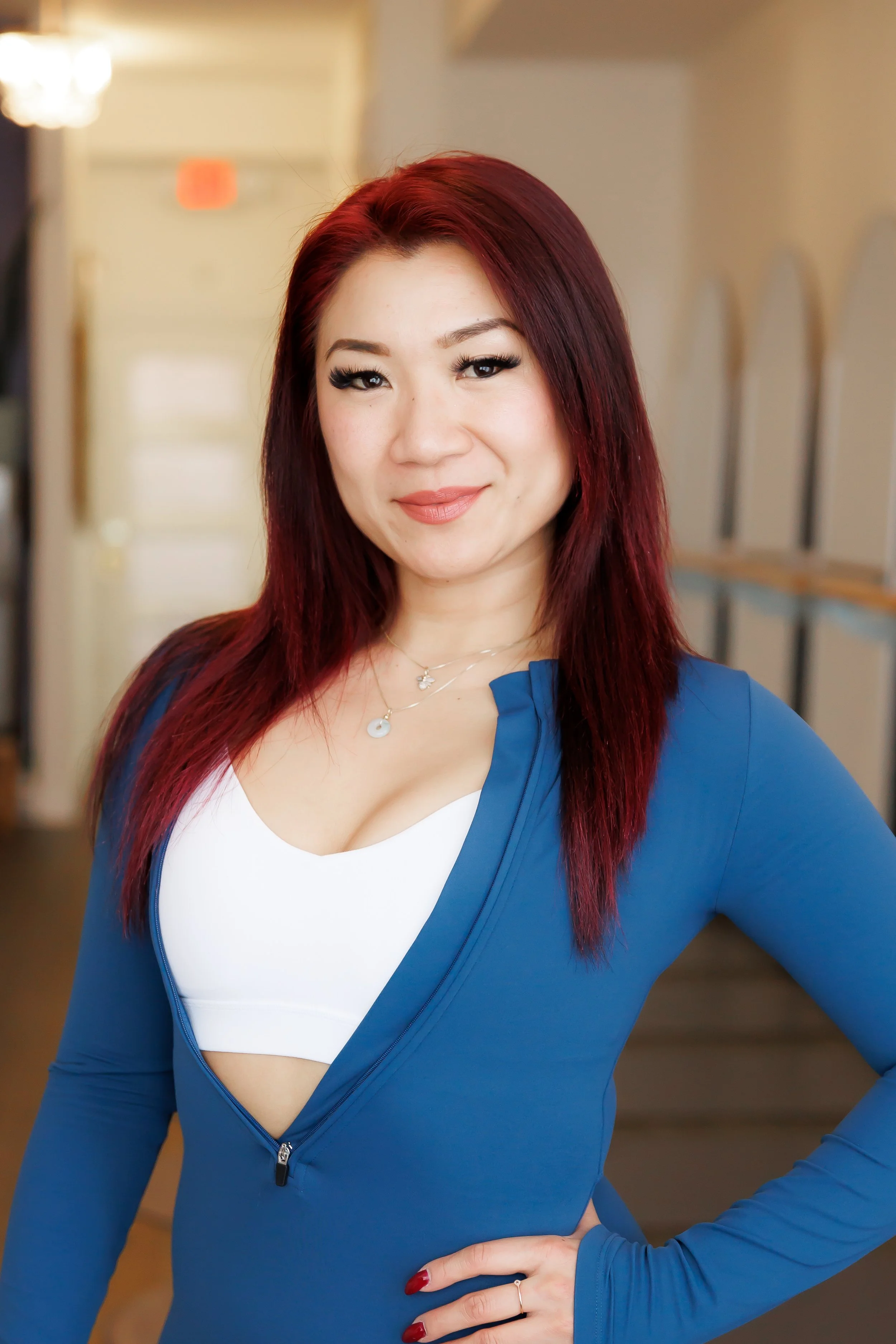 A woman with long red hair and light skin, smiling, wearing a blue zip-up athletic top over a white sports bra, standing indoors with a slightly blurred background. Pilates and barre studio in The Junction, Toronto.