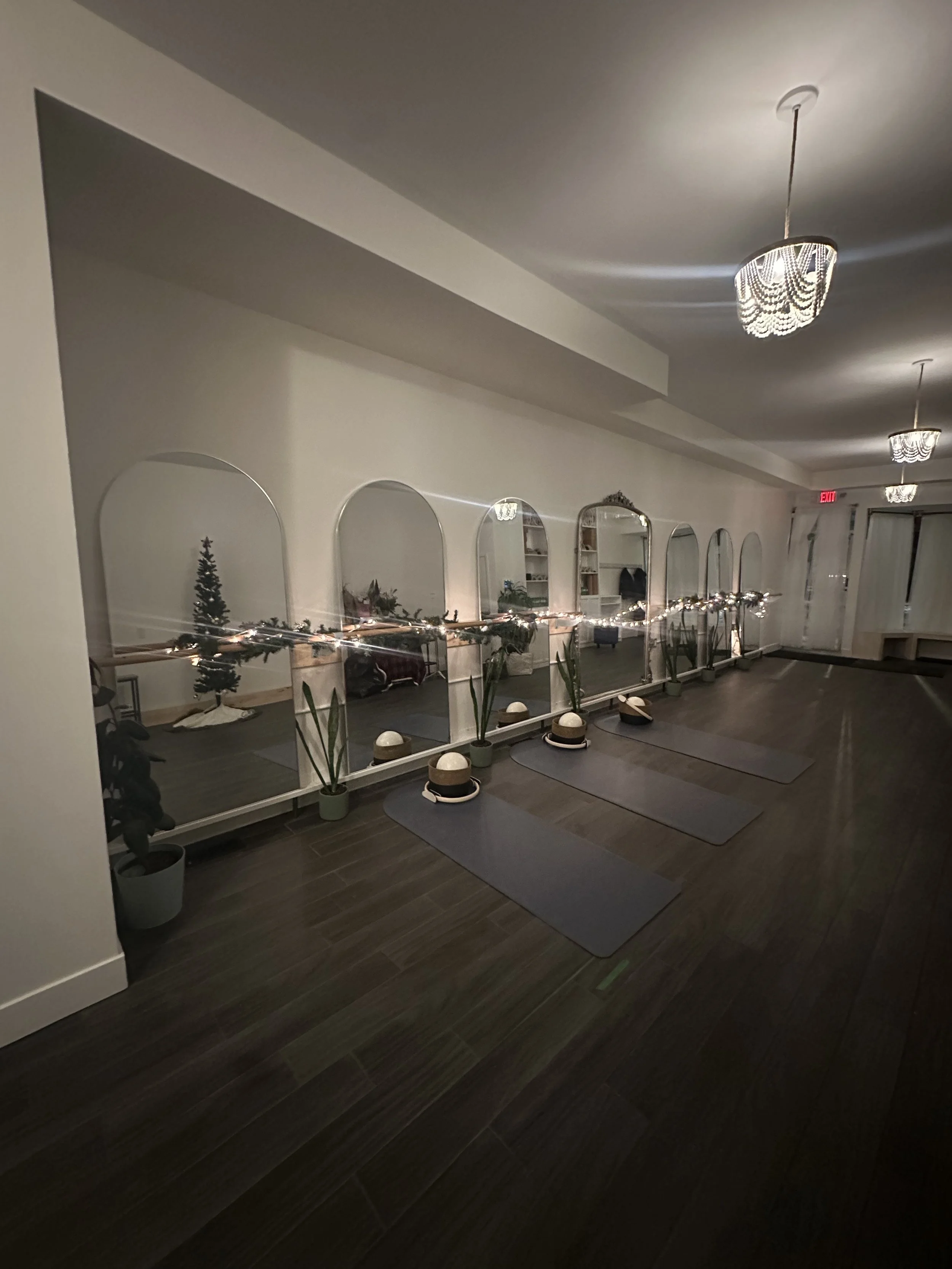 A row of yoga mats and helmets with headphones placed on the floor in front of six tall mirrors, decorated with small potted plants and string lights. The setting appears to be a wellness or pilates studio with dim lighting and ceiling chandeliers. P