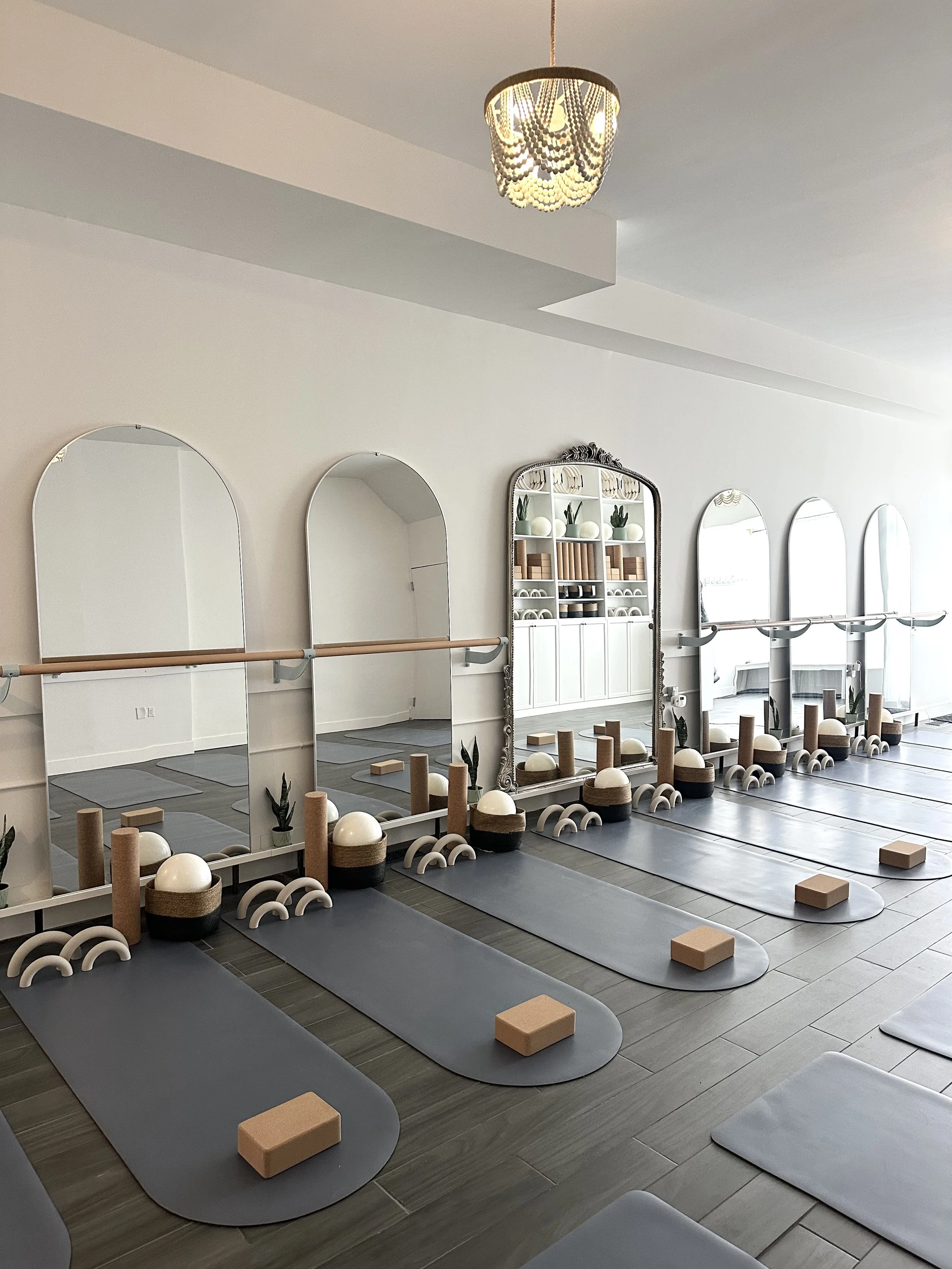 A yoga studio with four mats laid out on the floor, each with a yoga block, a small decorative arch, and a mirror behind it. The room has white walls, wooden flooring, and a modern chandelier hanging from the ceiling. Pilates and barre studio toronto