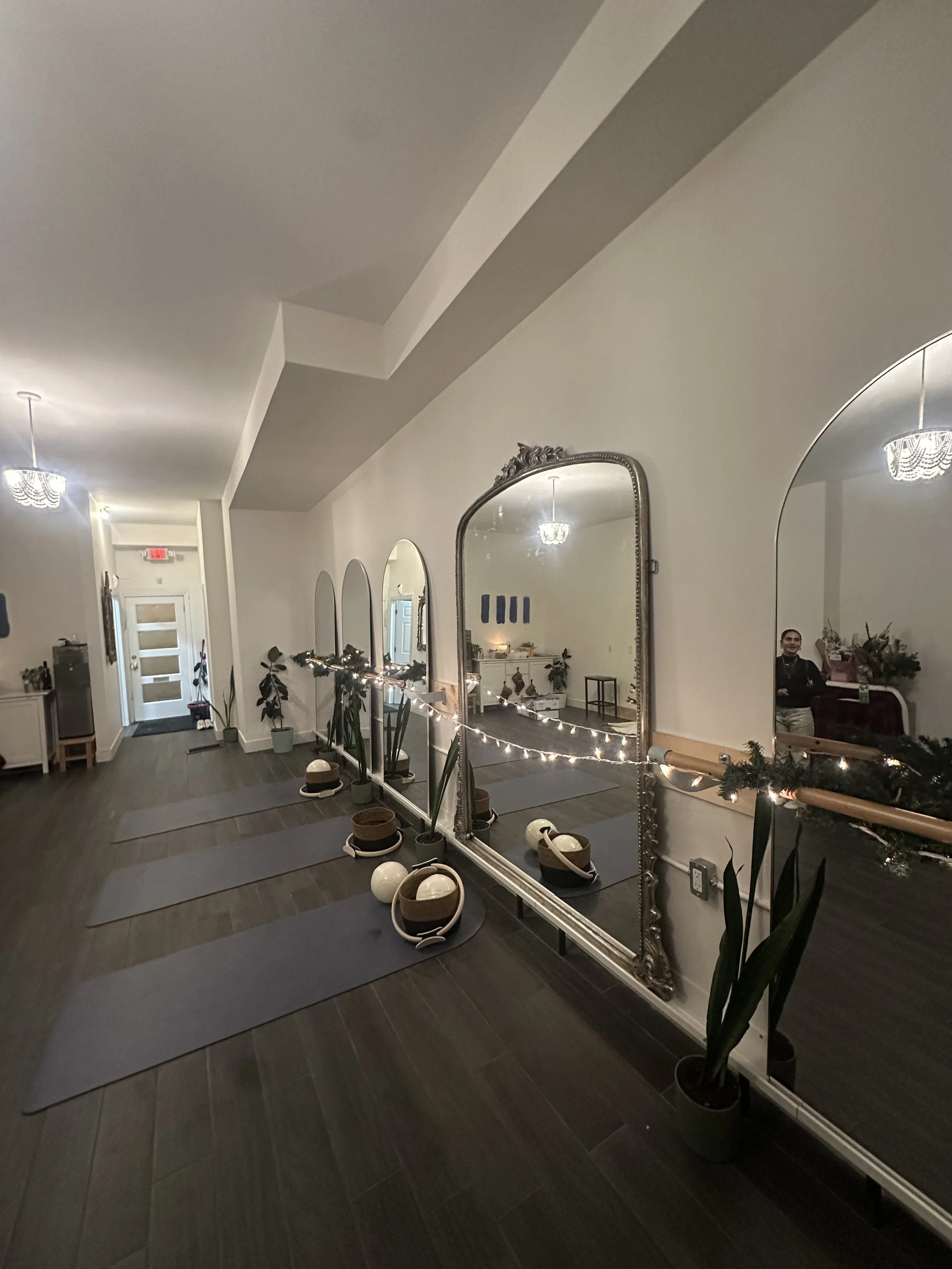 Interior of a room decorated for yoga with mirrors, plants, yoga mats, and ornaments, illuminated by chandeliers and string lights. Pilates and barre studio in The Junction, Toronto.
