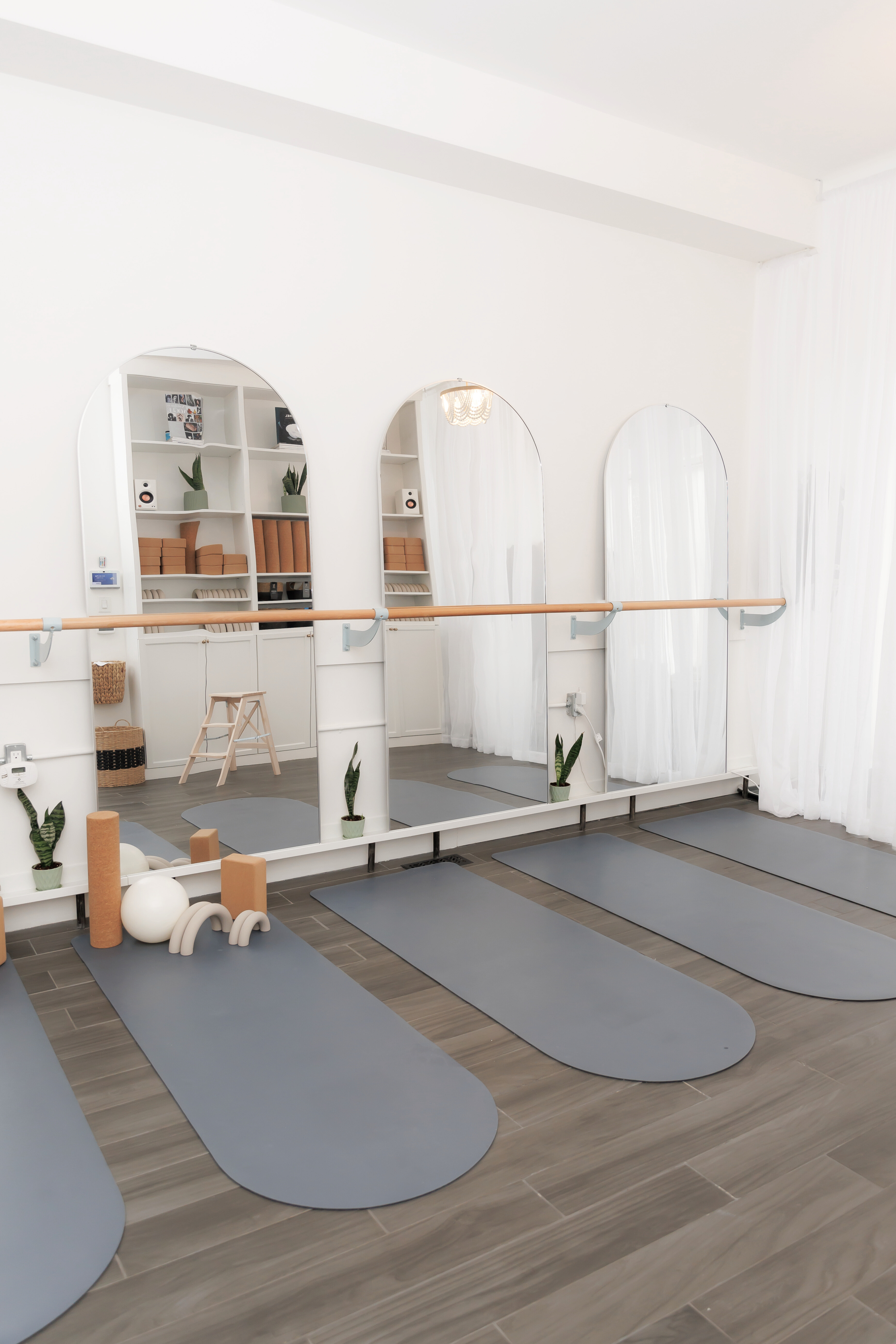 A modern, minimalistic yoga studio with four gray yoga mats placed on wooden floors, each mat in front of a mirror, with potted plants and yoga props like foam rollers and blocks. The studio has white walls, sheer curtains, and built-in shelves with books and small plants.