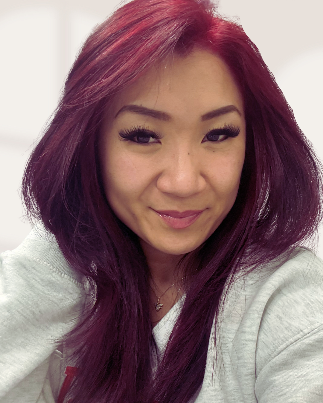 Close-up of a woman with long, wavy, reddish-purple hair, wearing makeup, a light-colored top, and a necklace, smiling softly at the camera. Pilates and barre studio in The Junction, Toronto.