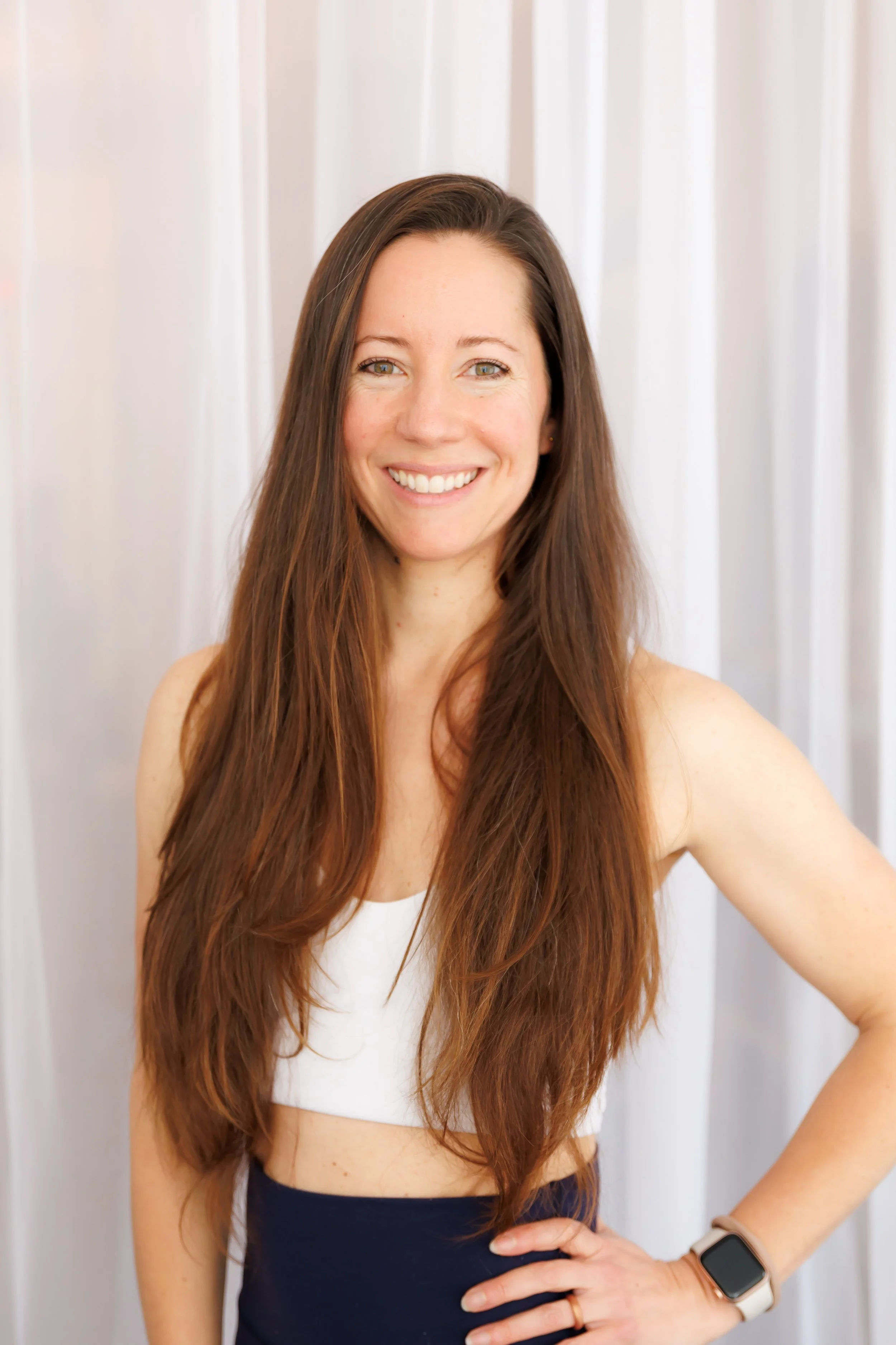 A woman with long brown hair smiling and wearing a white sleeveless top and black skirt, with a smartwatch on her wrist, standing in front of a light-colored curtain. Pilates and barre studio in The Junction, Toronto.