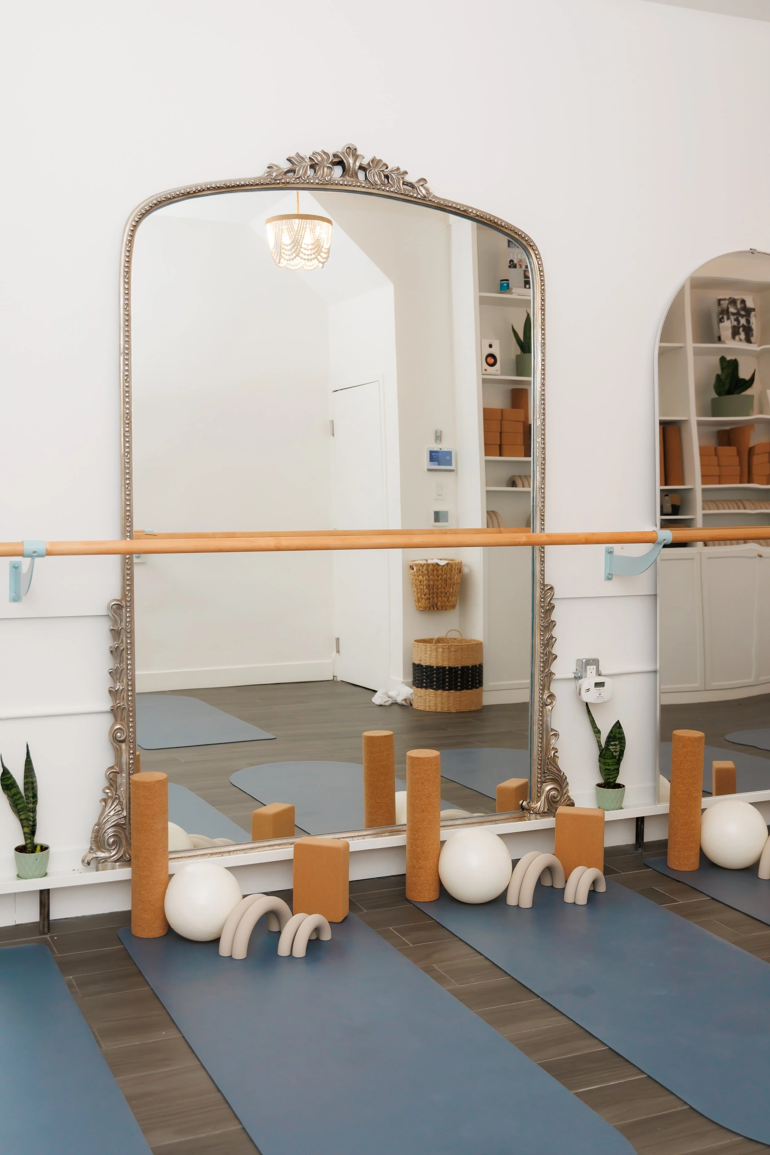Yoga studio with blue mats, white exercise balls, foam rollers, and small arches for practice, featuring large ornate mirror, plants, and shelves in background. Pilates and barre studio in The Junction, Toronto.