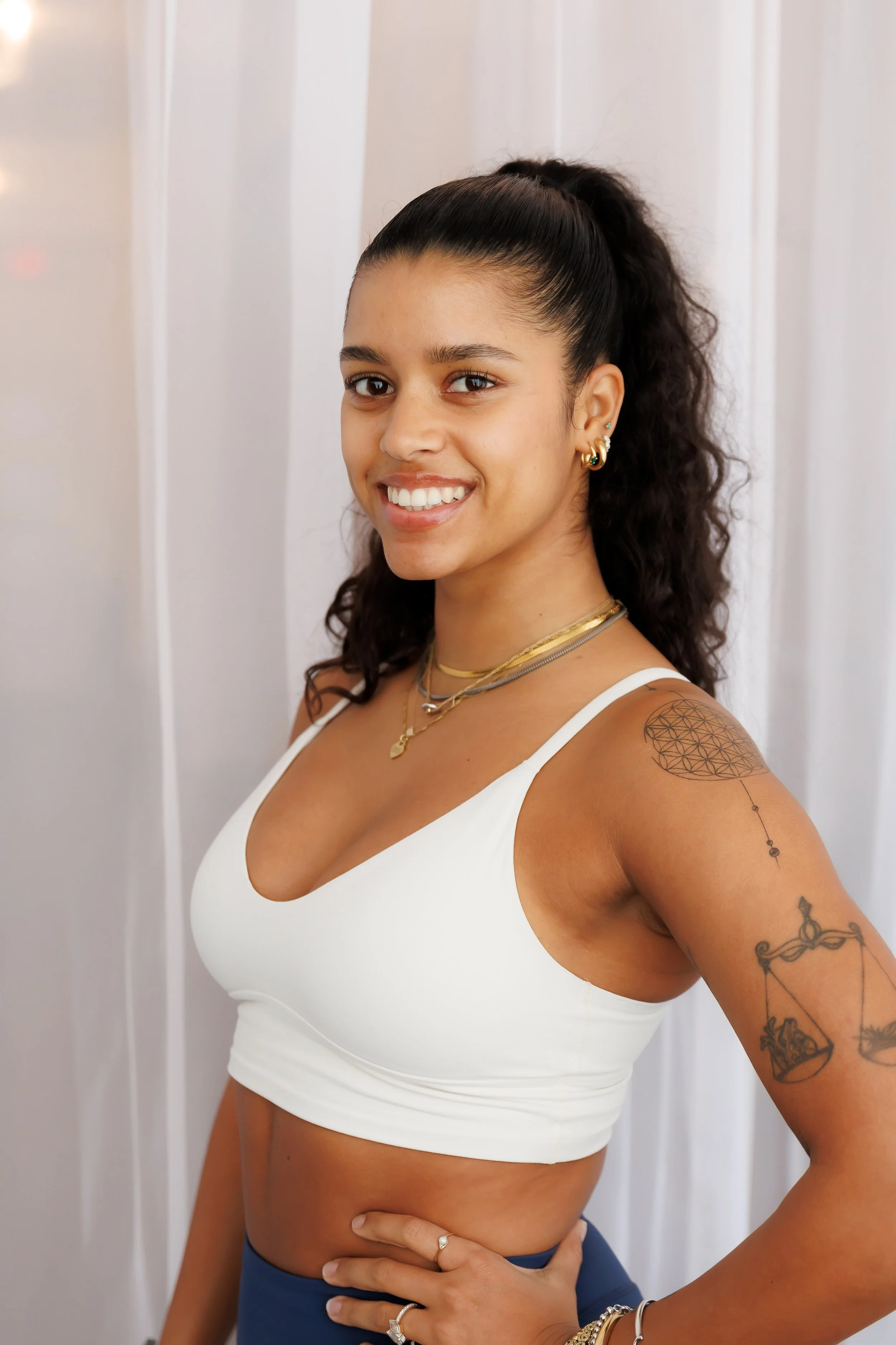 A young woman with dark, curly hair tied in a high ponytail, smiling and wearing a white sports bra, blue bottoms, and layered gold jewelry. She has tattoos on her left arm and stands in front of a white curtain. Pilates and barre studio in The Junct