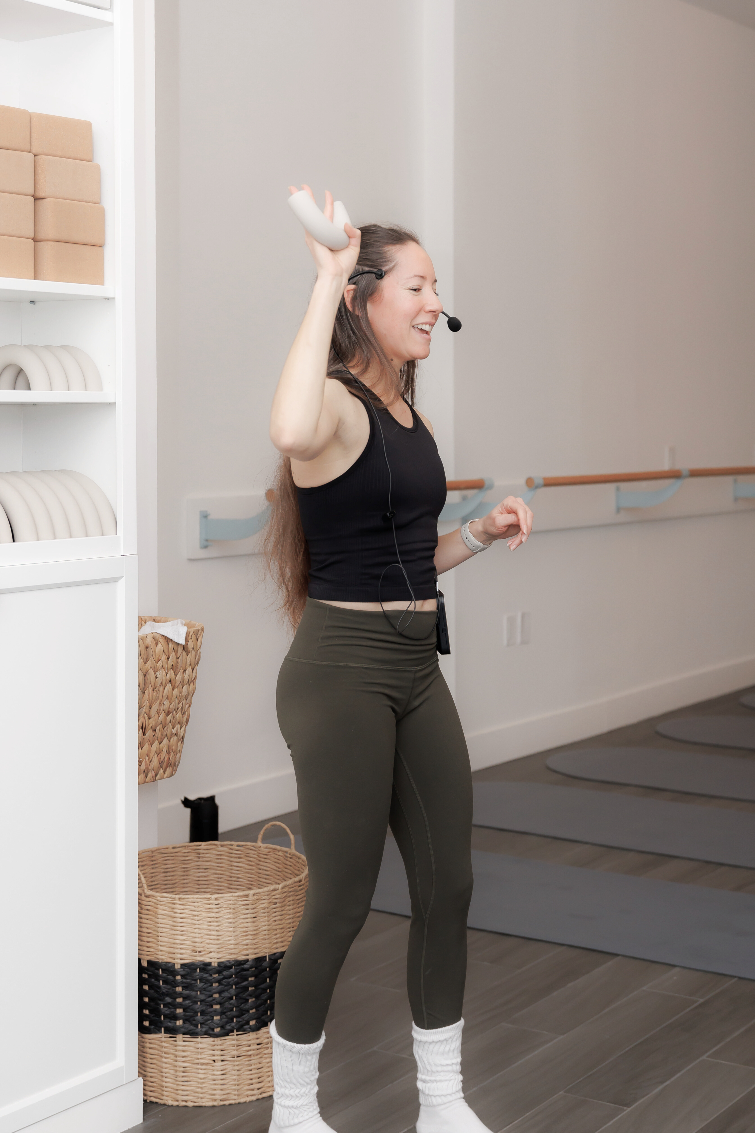 A woman leading a fitness or dance class, wearing athletic attire and a headset microphone, smiling and raising her right hand. Pilates and barre studio in The Junction, Toronto.