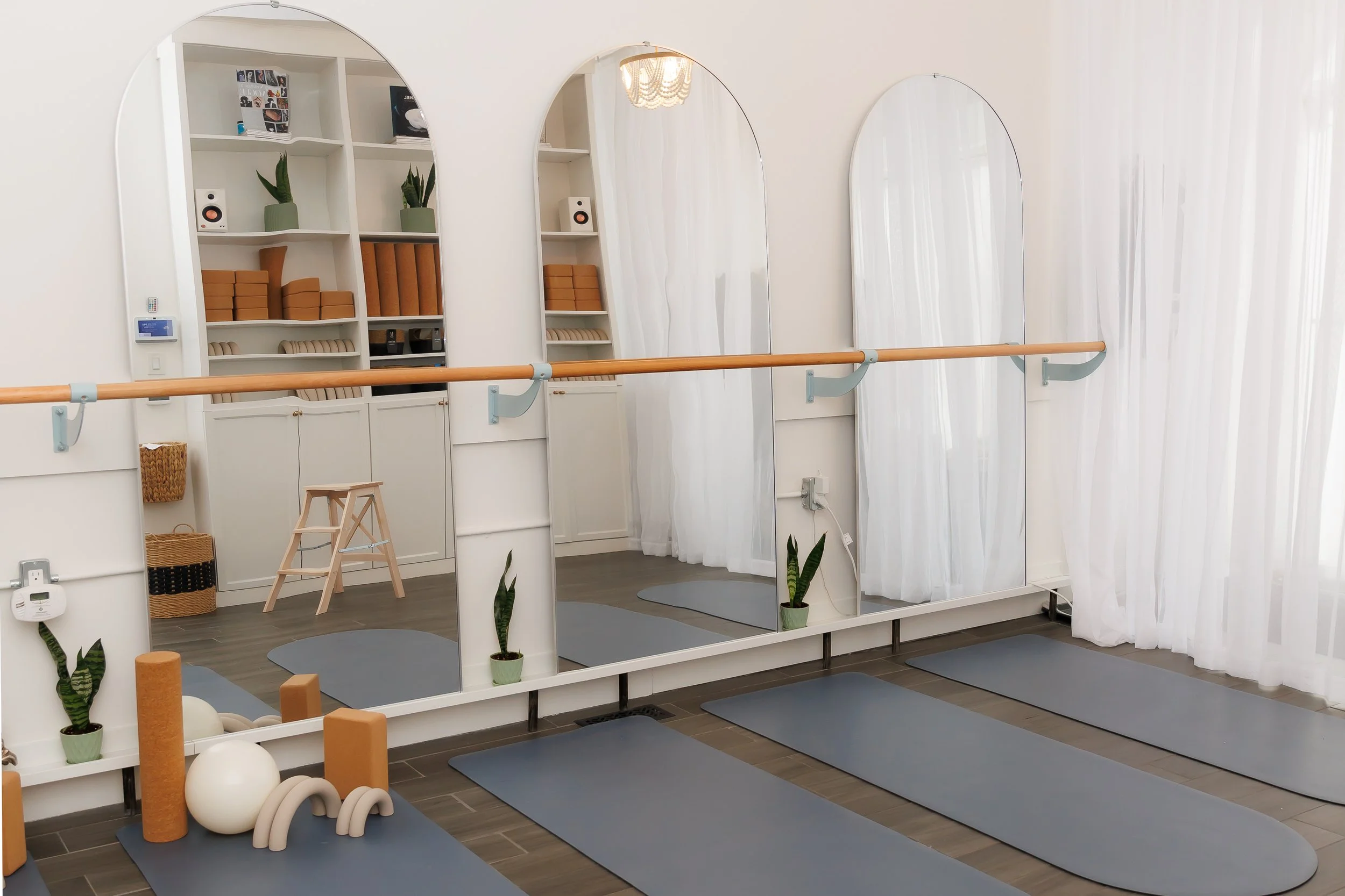 Yoga studio with four gray mats on wooden floor, mirrored wall, ballet barre, white curtains, green potted plants, and yoga props. Pilates and barre studio in The Junction, Toronto.