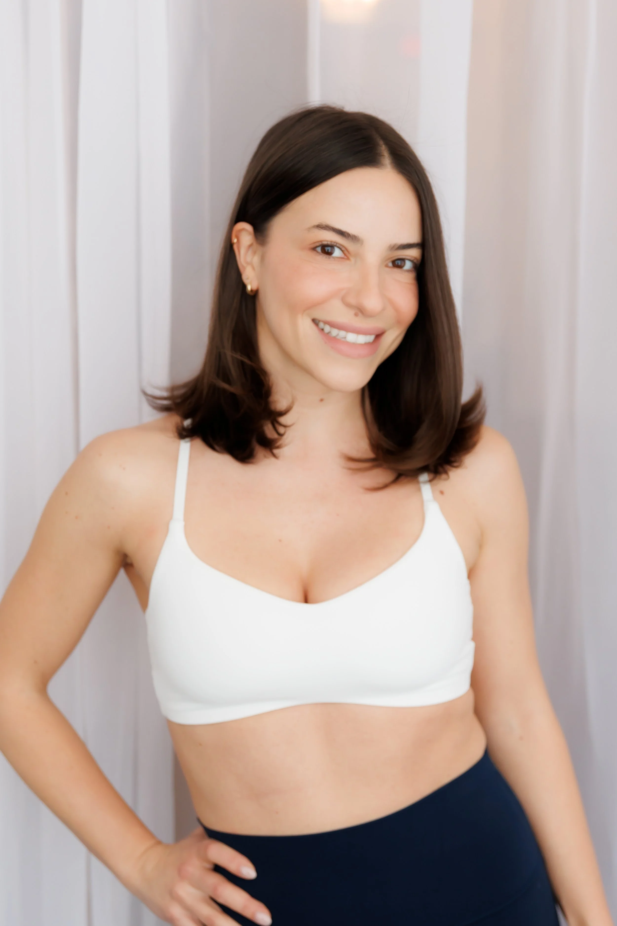 A woman with shoulder-length brown hair, smiling, wearing a white tank top and black pants, standing in front of a light-colored wall. Pilates and barre studio in The Junction, Toronto.