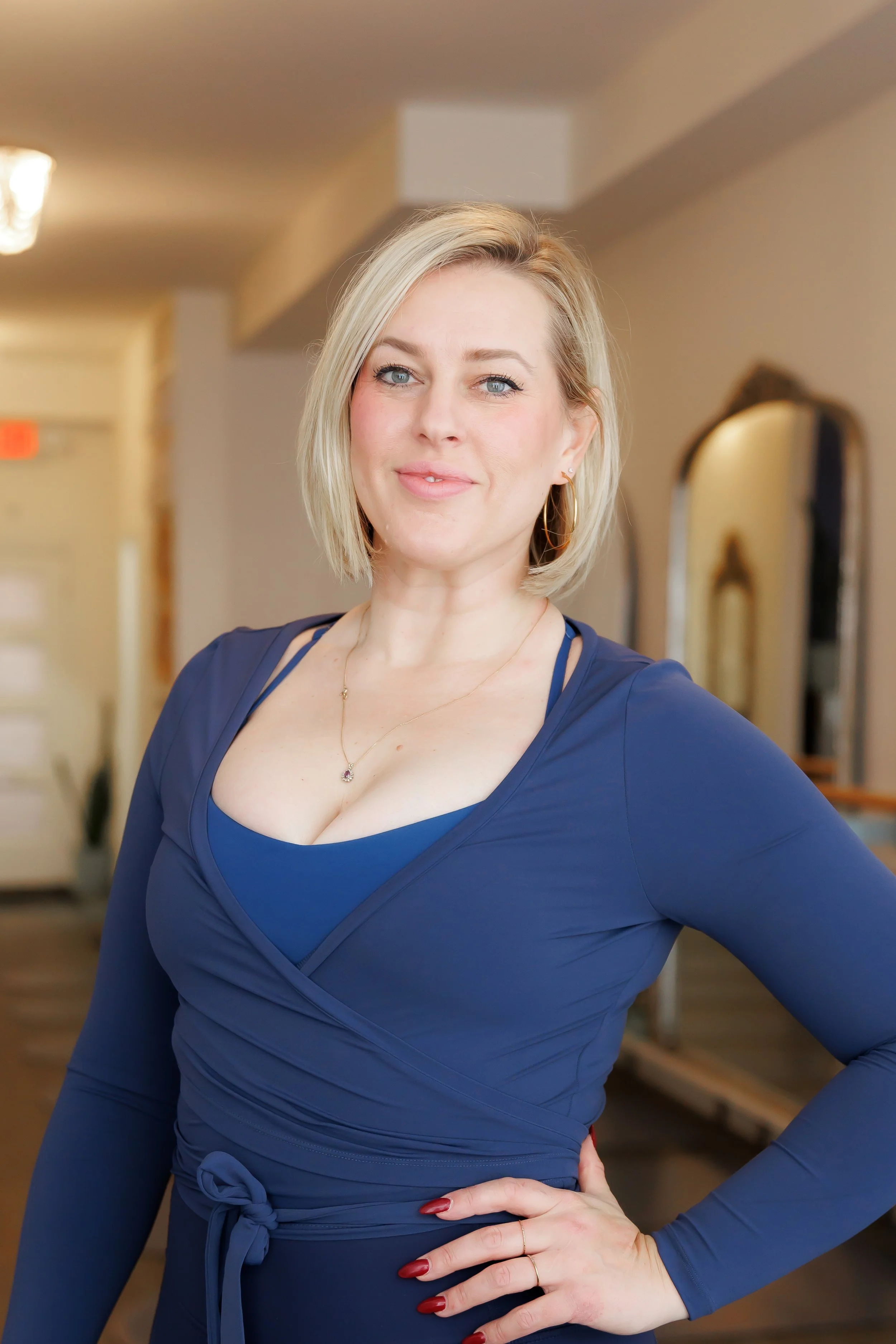 A woman with shoulder-length blonde hair, wearing a navy blue dress with a matching top underneath, standing in an indoor setting with a mirror and door in the background, smiling softly. Pilates and barre studio in The Junction, Toronto.
