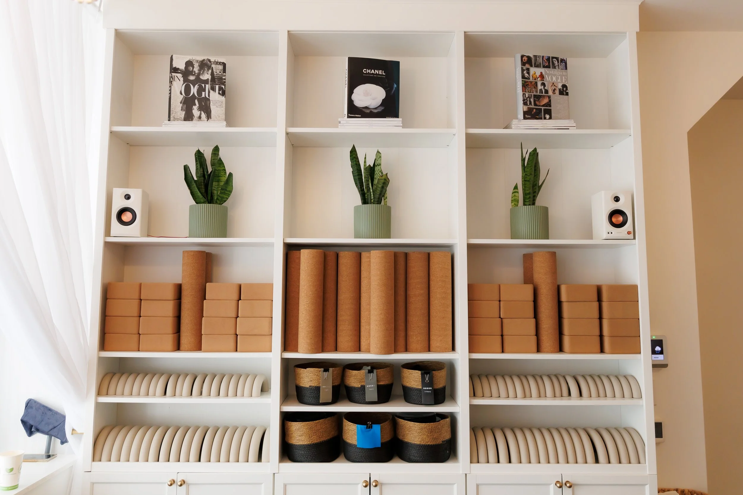 A white bookshelf containing books, potted plants, speakers, and storage boxes in a well-lit room. Pilates and barre studio in The Junction, Toronto.