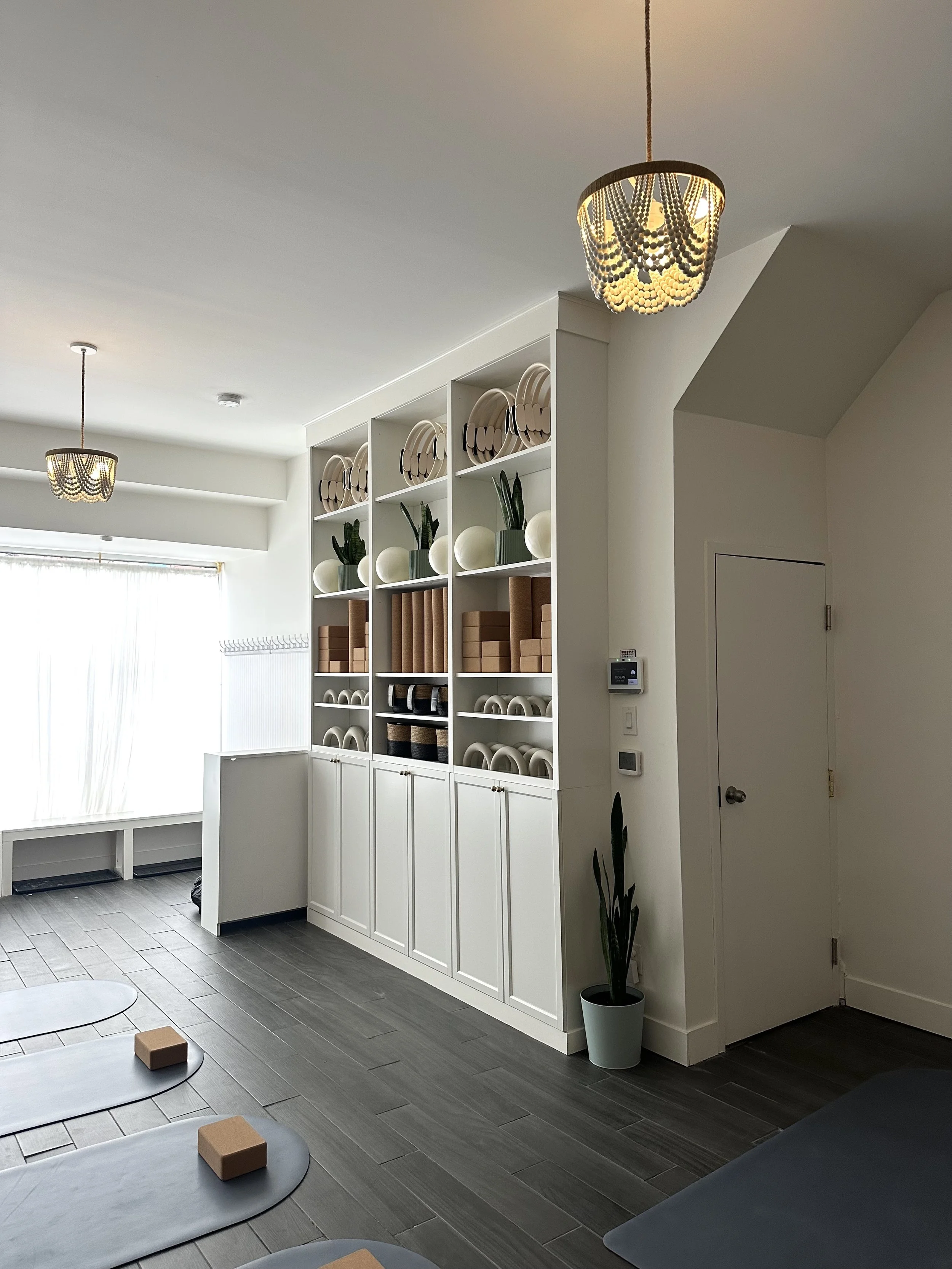 Interior of a room with white shelves holding yoga props, potted plants, and decorative objects. The room has gray flooring, two gold pendant lights, and natural light coming through a window. Pilates and barre studio in The Junction, Toronto.