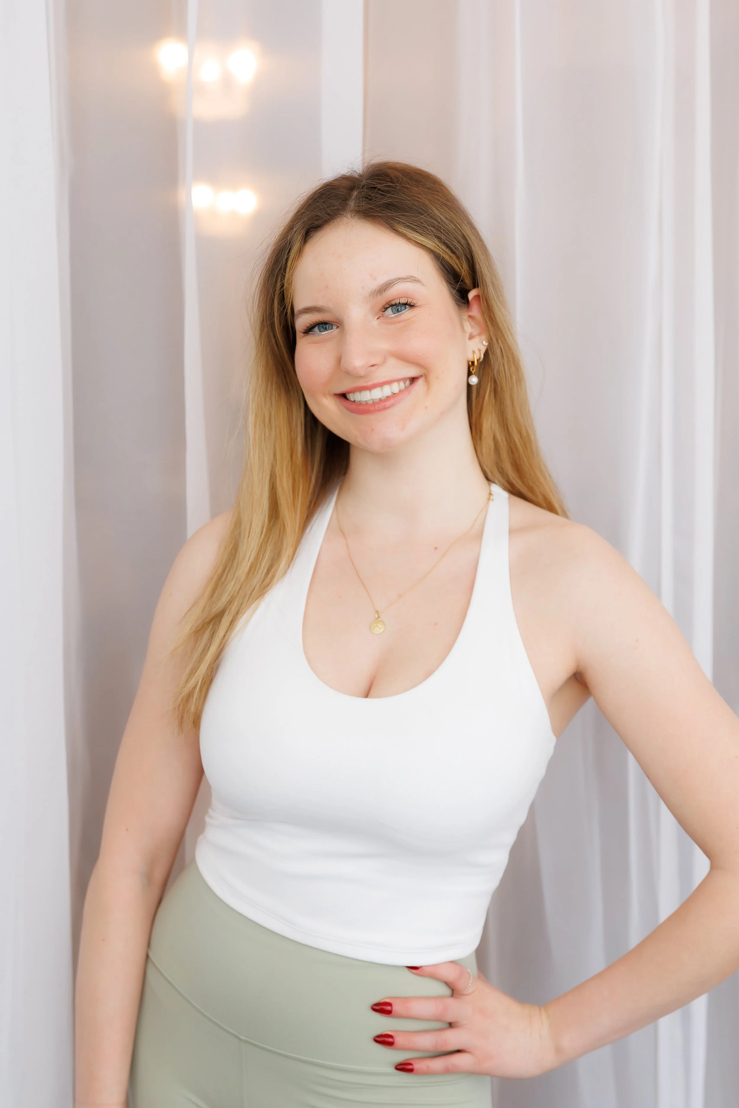 A young woman with long, light brown hair, blue eyes, and fair skin, smiling at the camera. She is wearing a white tank top, light-colored pants, and gold jewelry, including a necklace and earrings. She stands indoors against a white background with soft lighting.