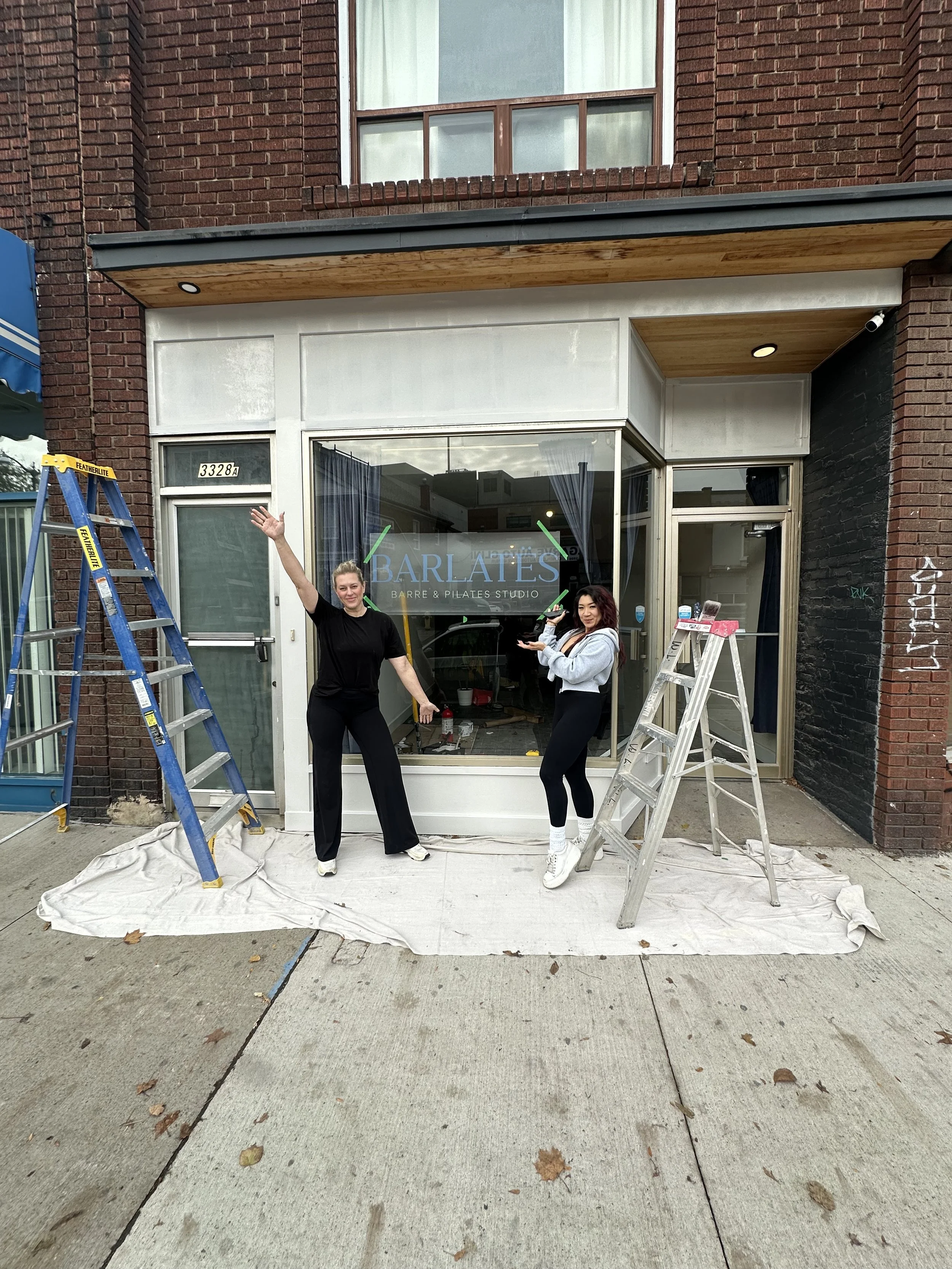 Two women standing outside a storefront with the sign 'BARLATAS' that reads 'Barre & Pilates Studio.' They are holding paint rollers, and there are two ladders on either side, with a white cloth spread on the sidewalk beneath them. The storefront app