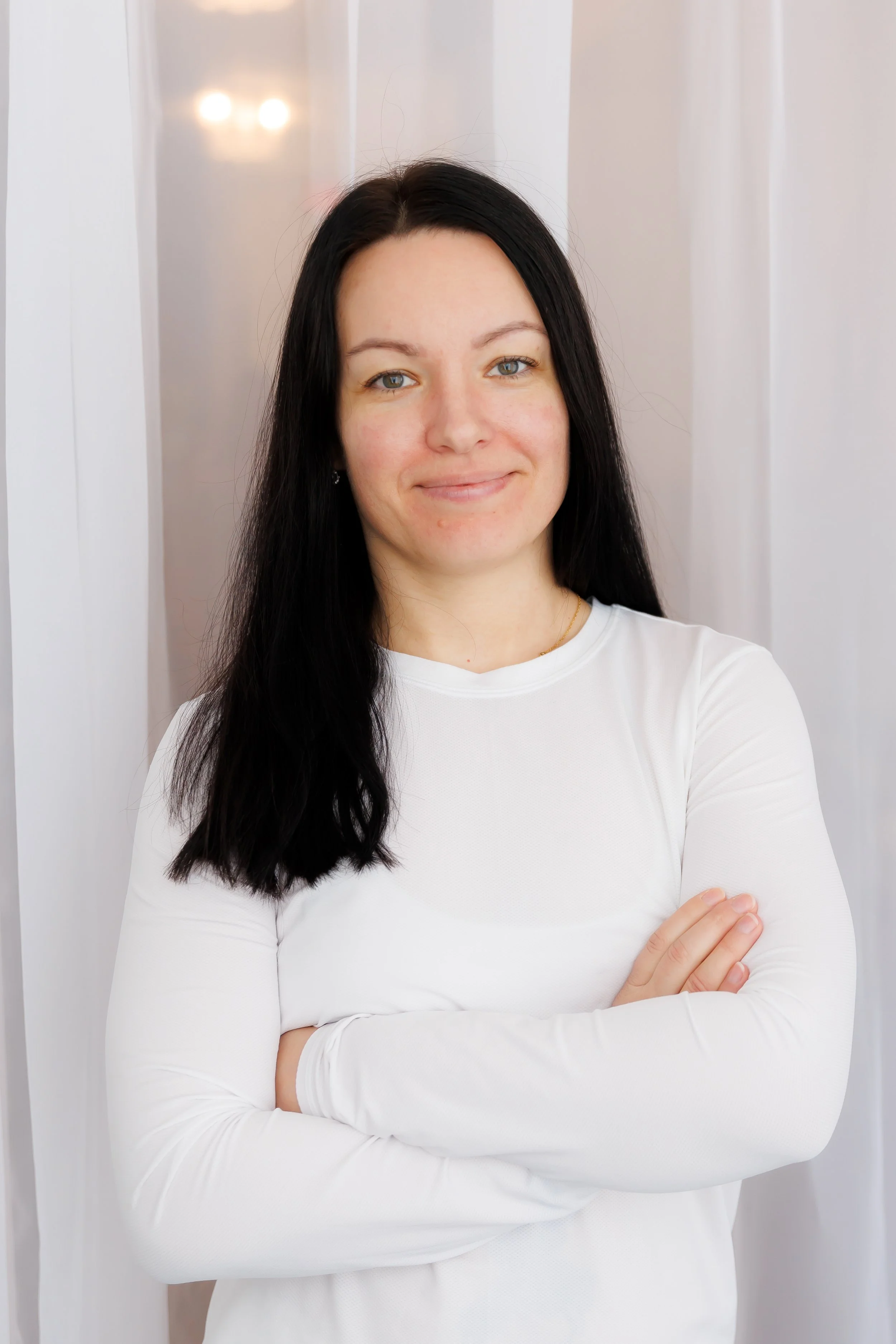 A woman with long black hair, wearing a white long-sleeve shirt, standing with arms crossed in front of a white curtain backdrop, smiling gently at the camera. Pilates and barre studio in The Junction, Toronto.