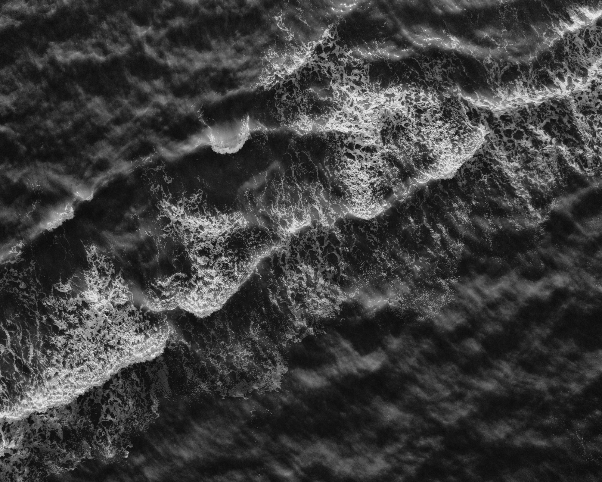 Aerial black and white photograph of ocean waves crashing on the shoreline.