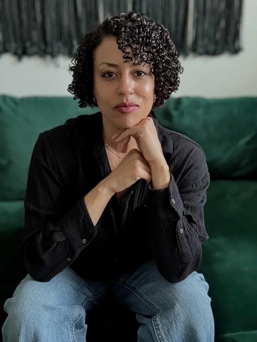 A mixed-race woman with short curly hair, wearing a black long-sleeve shirt and denim jeans sitting on a green couch with a dark wall hanging behind her.