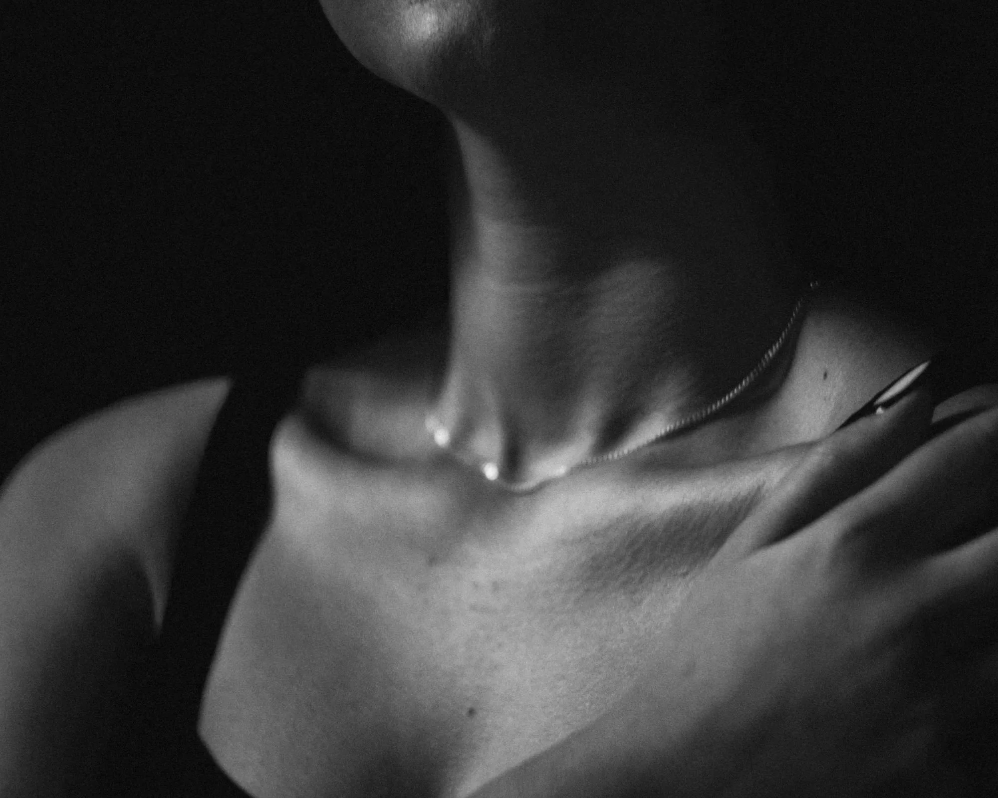Close-up of a woman's neck and shoulder, with her hand resting on her collarbone, wearing a delicate necklace and black sleeveless top.
