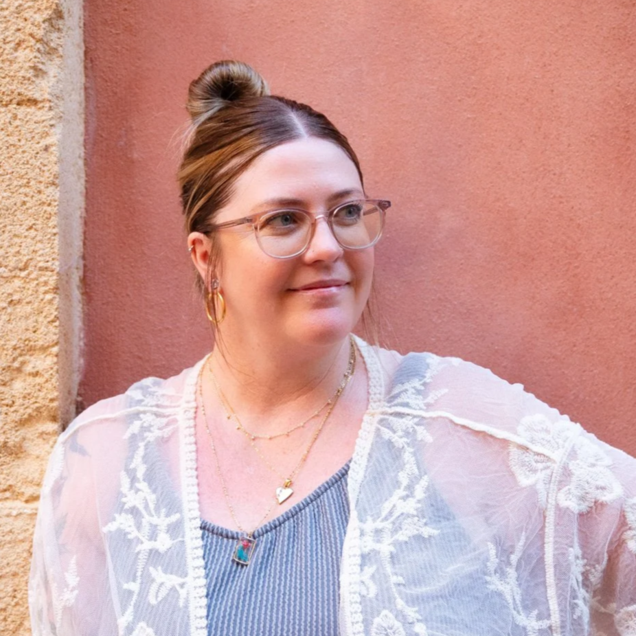 A woman with glasses and jewelry stands against a pink and beige wall.