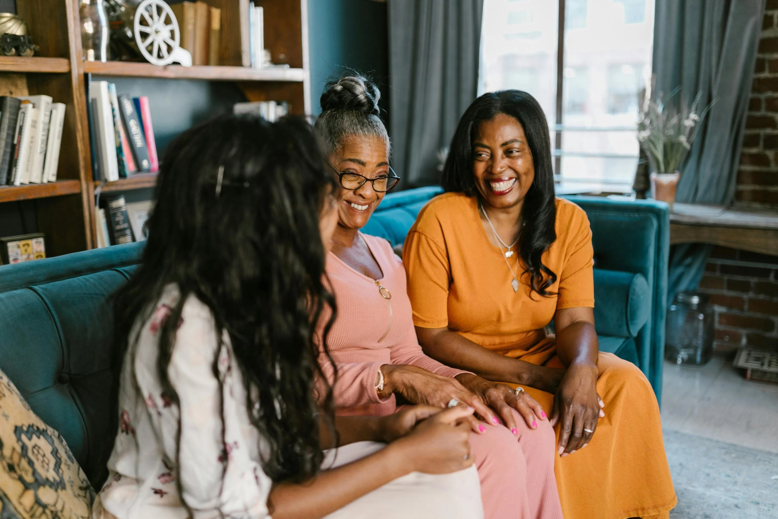 Three women sitting on a green sofa, smiling and talking in a cozy home with a bookshelf and houseplant in the background.