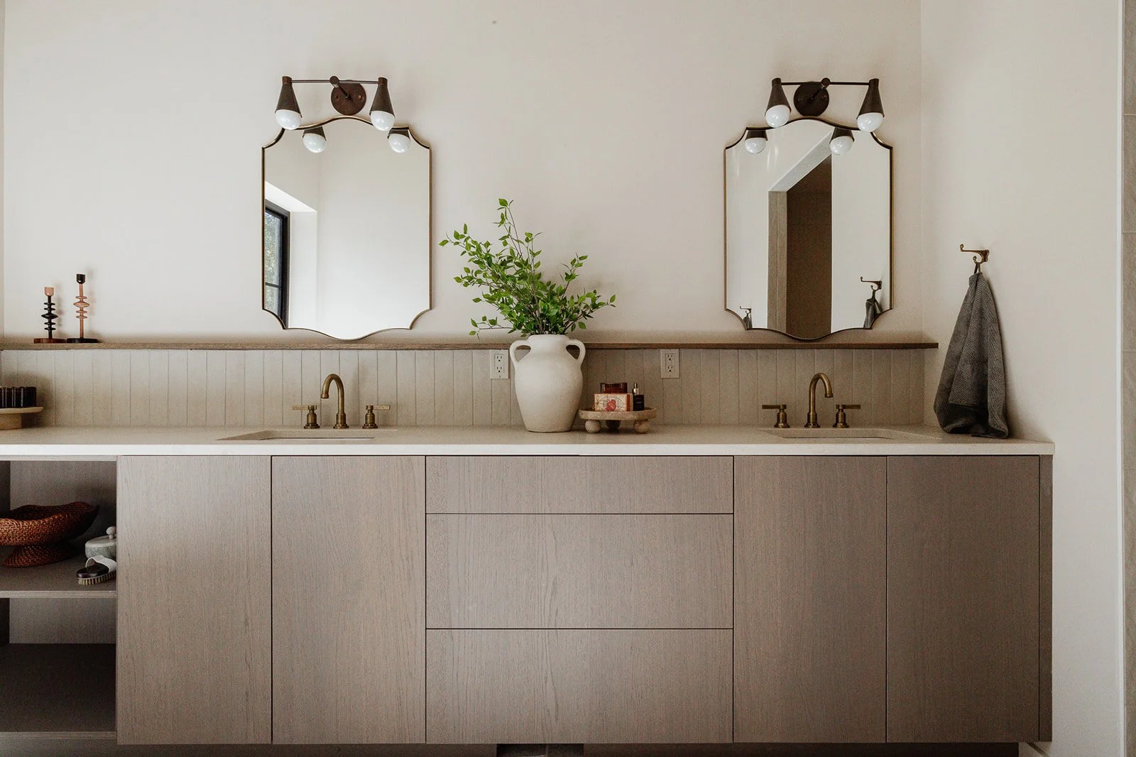 Double bathroom vanity with two mirrors, brass faucets, a plant in a white vase, and decorative items. Beige cabinets and a beige backsplash.