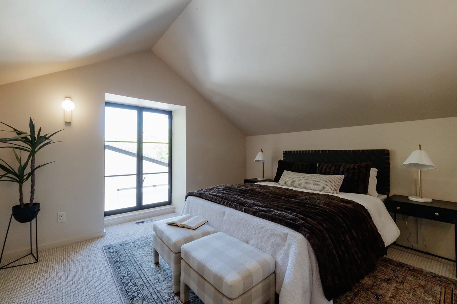 A cozy bedroom with a sloped ceiling, featuring a large bed with a dark headboard, white bedding, and a dark throw blanket, flanked by two nightstands with matching lamps, a potted plant, and a sliding glass door to a balcony.