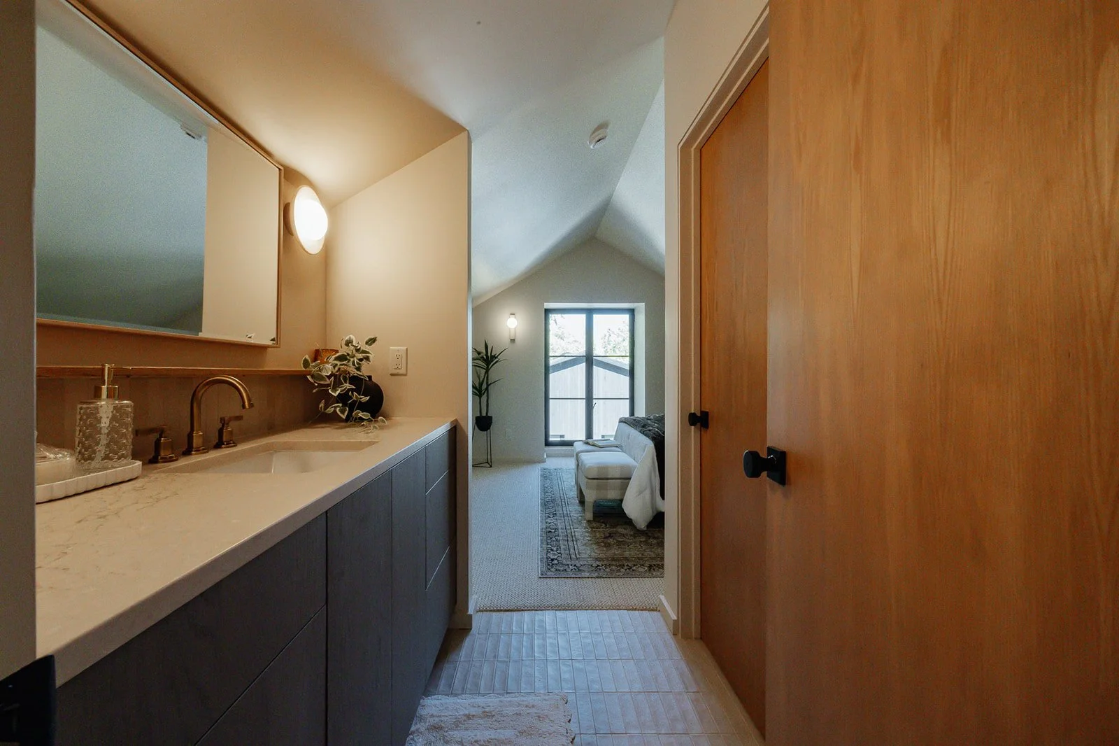 View of a cozy living area seen from a bathroom with a sink and mirror. The room has a sloped ceiling, a window, a sofa, and indoor plants.