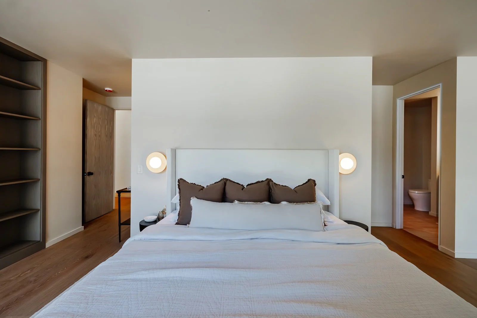 Modern bedroom with a large bed, white headboard, and multiple pillows. Nightstands with lamps on both sides, a wooden shelf to the left, and a doorway to the bathroom to the right.