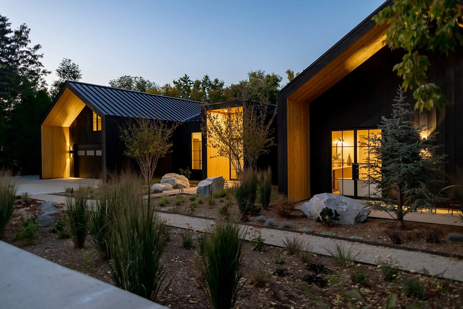 Modern houses with black exteriors, large windows, and warm interior lighting at dusk, surrounded by landscaped yard with rocks and small trees.