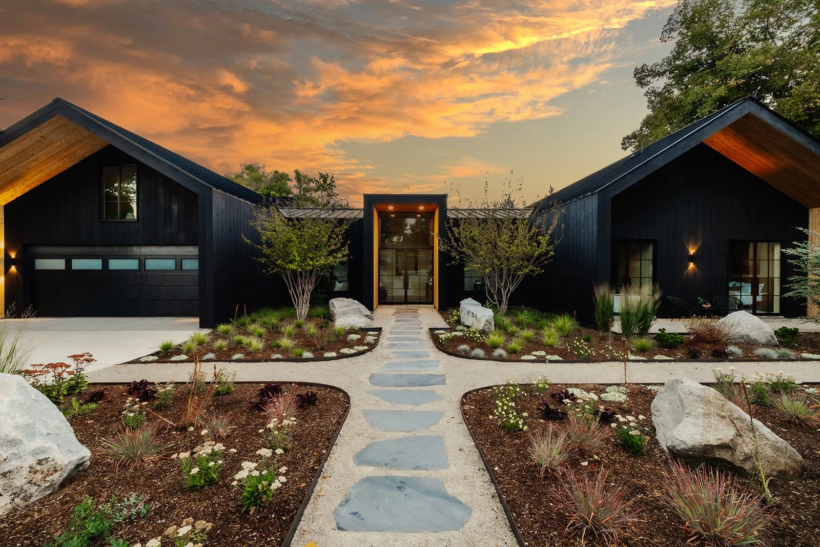 Modern house with black exterior and large glass entrance, landscaped front yard with rocks and plants, sunset sky with orange and pink clouds.
