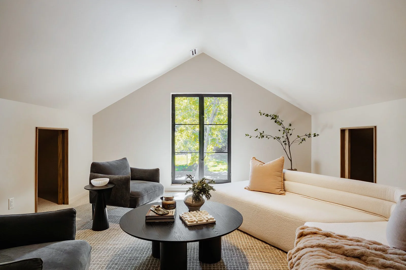 Bright living room with a large window, white walls, and comfortable seating including a long beige sofa, a black armchair, and a dark gray armchair. There is a round black coffee table with decorative items, a small side table, and a potted plant ne