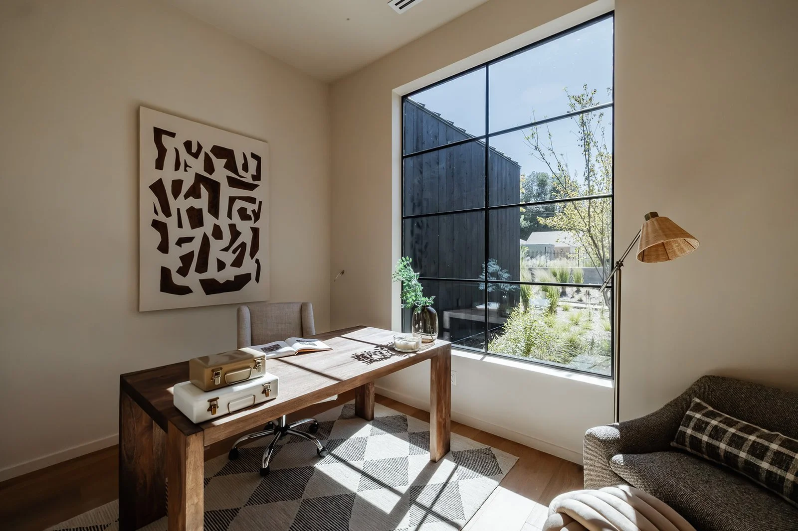 A bright, minimalist home office with a large window, a wooden desk, a beige chair, a patterned rug, decorative art on the wall, and a gray armchair with a plaid pillow.