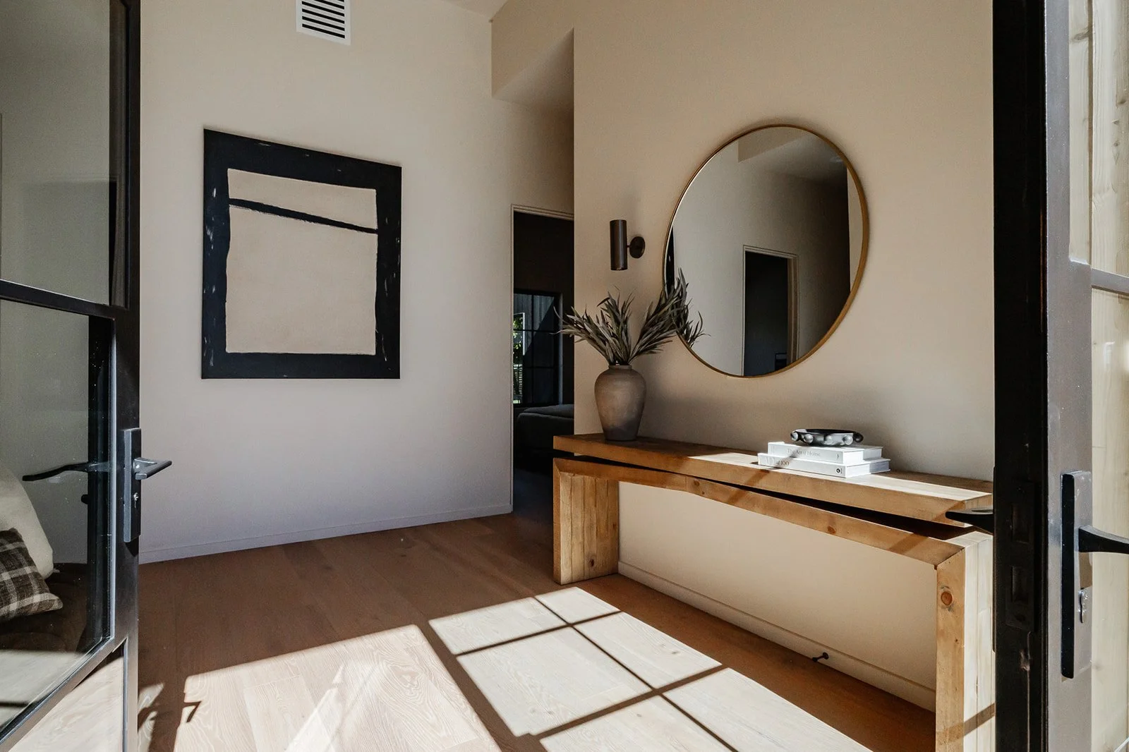 A modern entryway with a wooden console table, a round mirror, a large ceramic vase with dried plants, and abstract wall art. Sunlight creates a grid shadow pattern on the wooden floor.