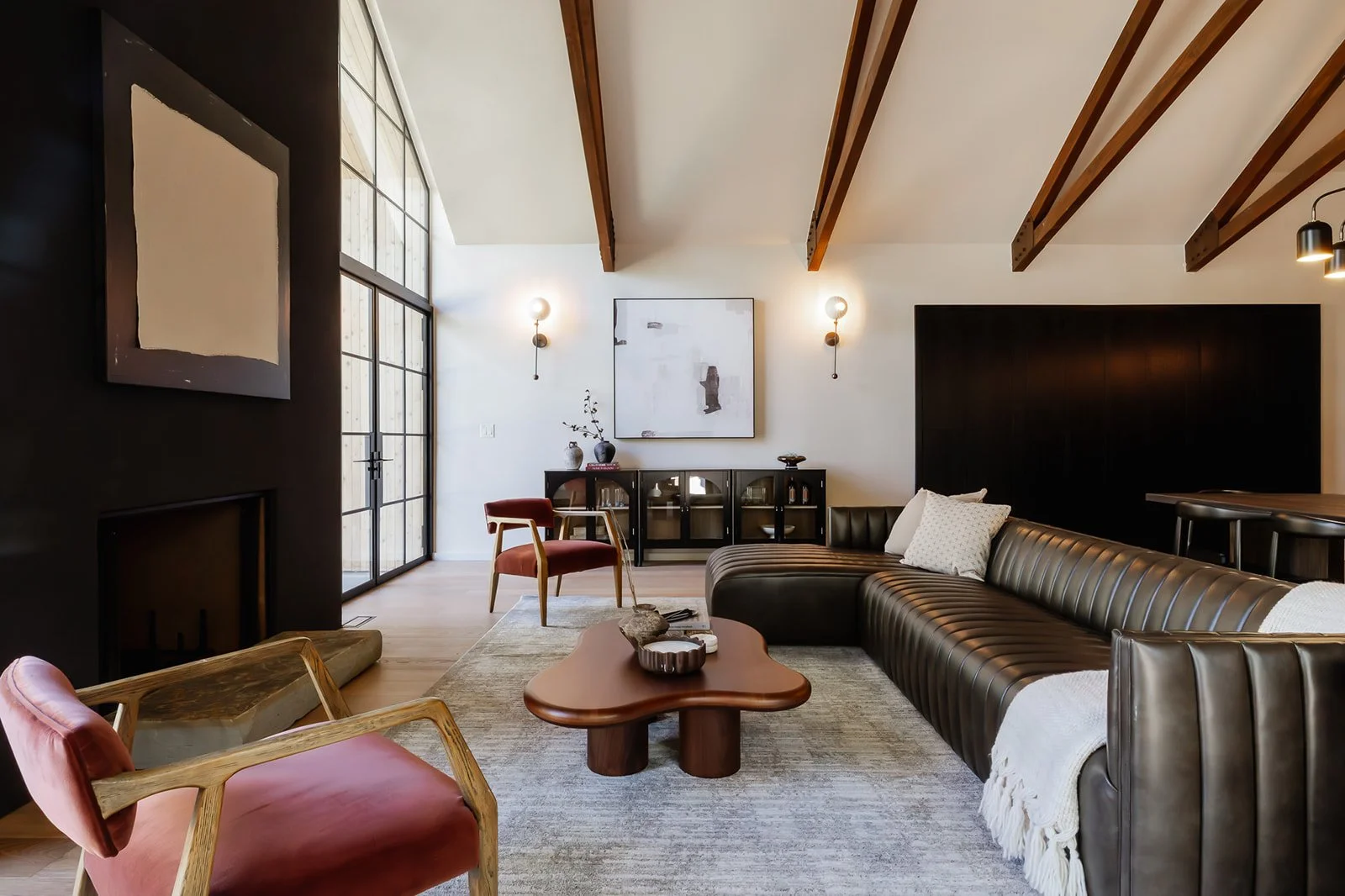 Modern living room with a large black leather sectional sofa, a wooden coffee table, and a couple of pink and wooden armchairs. There is a dark accent wall, a light-colored rug, and a large window or door with glass panes. Decor includes abstract pai