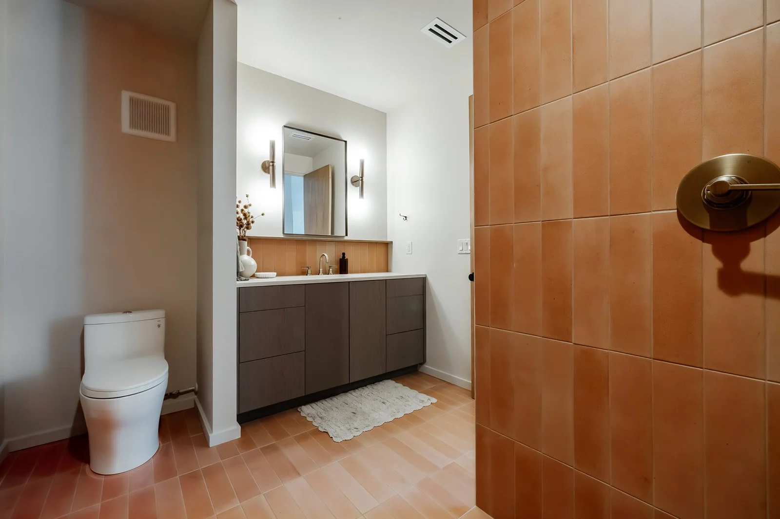 Modern bathroom with a white toilet, a gray vanity with a white countertop, a framed mirror, and decorative items including a vase with dried flowers and a bottle, illuminated by wall-mounted light fixtures.