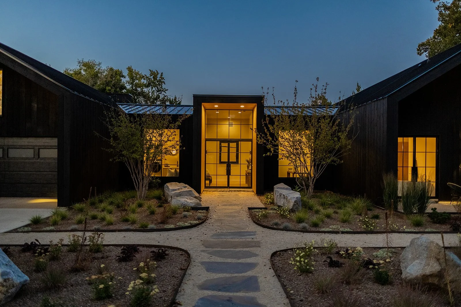 Modern house with black exterior, illuminated windows and front door, landscaped front yard with plants and rocks, and a stone pathway leading to the entrance, during dusk or early evening.