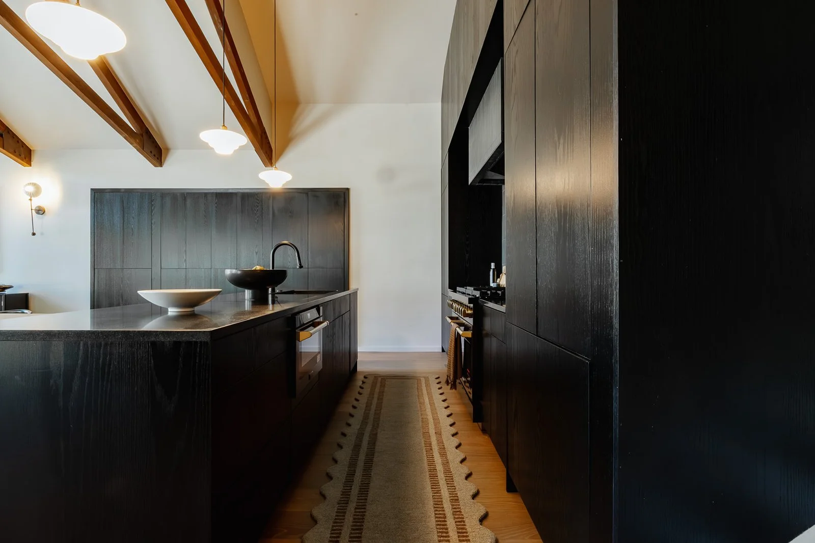 Modern kitchen with black cabinets, wood beams, and pendant lighting, featuring a kitchen island with two bowls and a wall-mounted oven, running along a wooden floor with a beige rug.