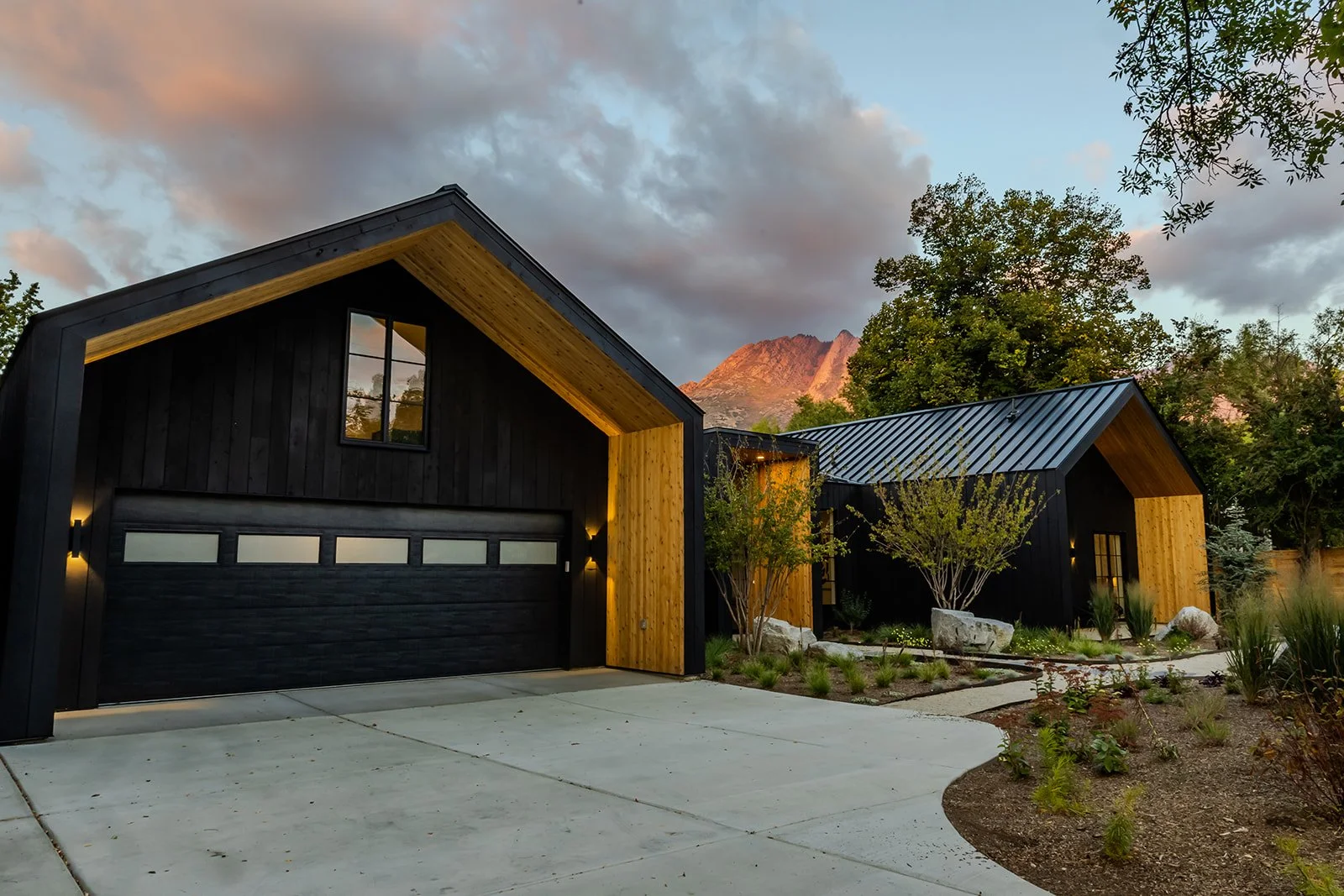 Modern black and wood houses with landscaped front yard, trees, and mountains in the background at sunset.