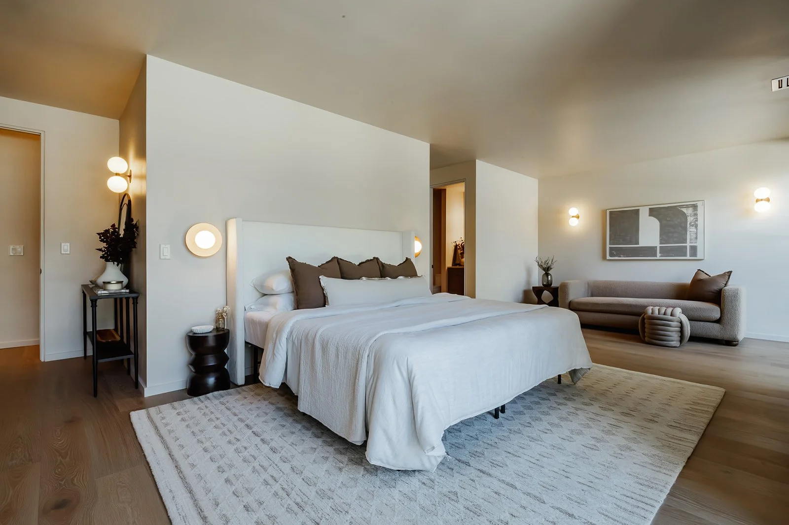 Modern bedroom with a large white bed, dark pillows, surrounding warm lighting, wooden floors, a sofa with cushions, framed artwork, and minimalistic decor.