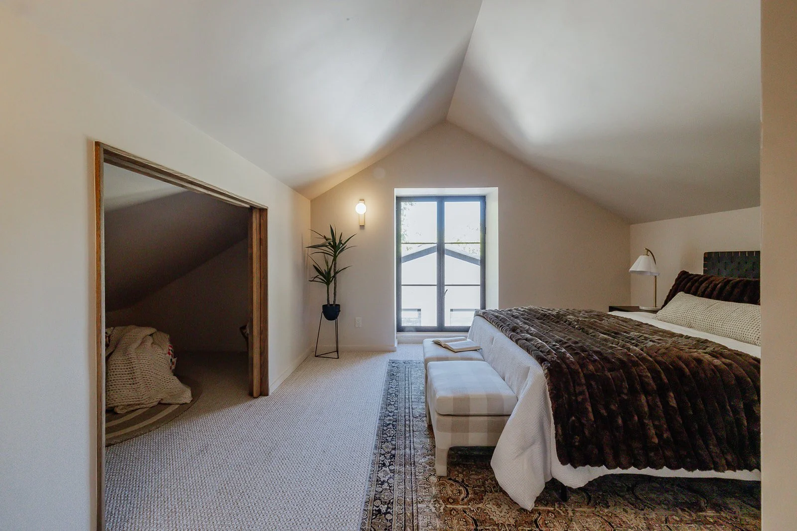 Bedroom with a large bed covered in a brown fur blanket, a window with black framing, a white bench at the foot of the bed, a small nightstand with a lamp, a tall plant next to the window, and a small room or closet to the left with a blanket on the
